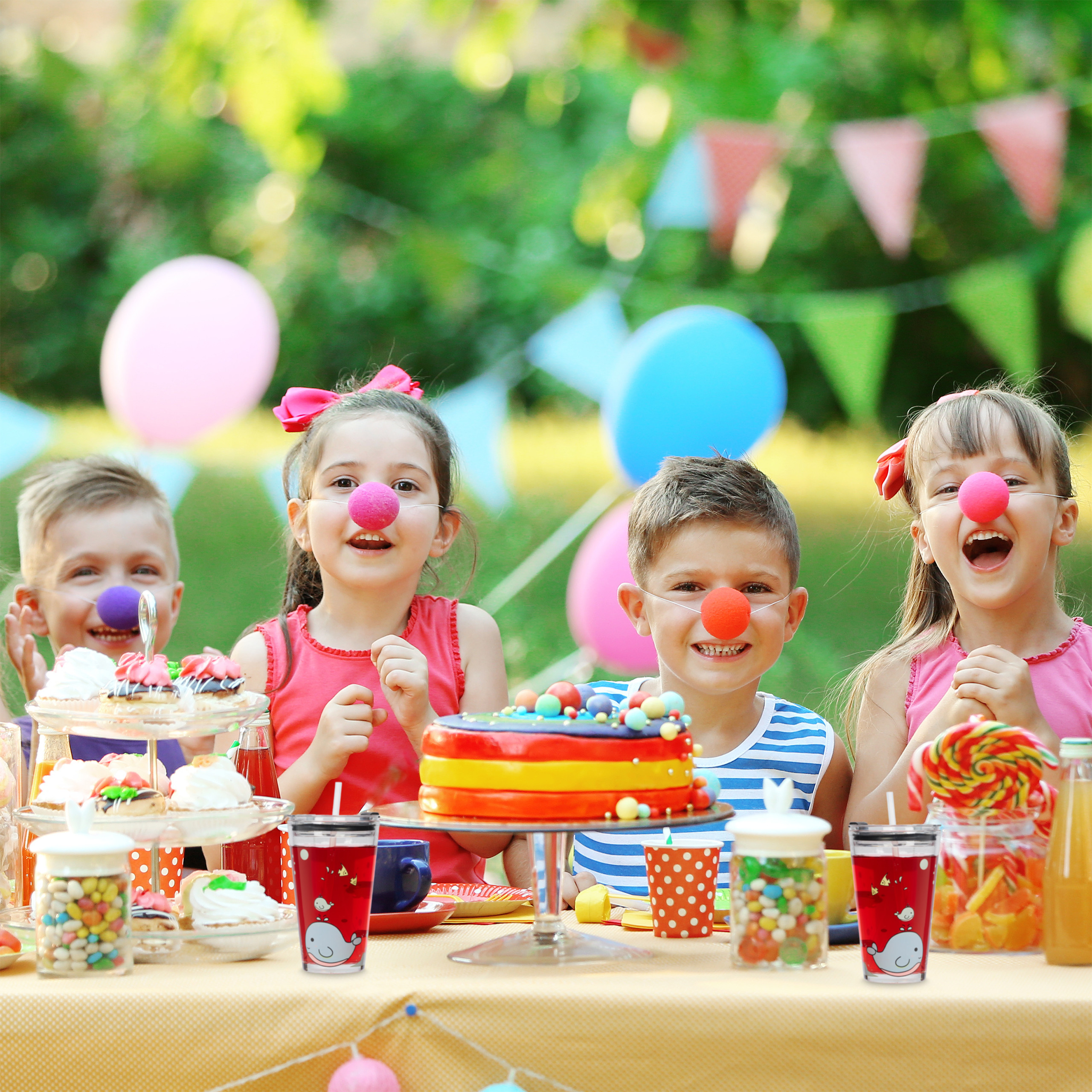 Vier Kinder mit Clownsnasen lächeln an einem Partytisch mit Kuchen und Süßigkeiten im Freien.