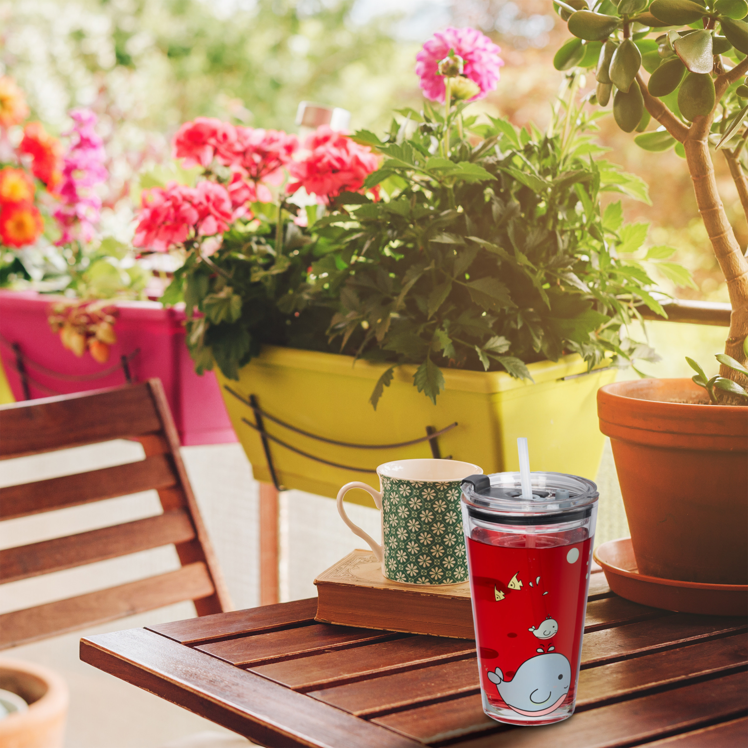 Ein Becher mit rotem Getränk, eine gemusterte Tasse auf einem Buch und Pflanzen auf einem sonnigen Holztisch.