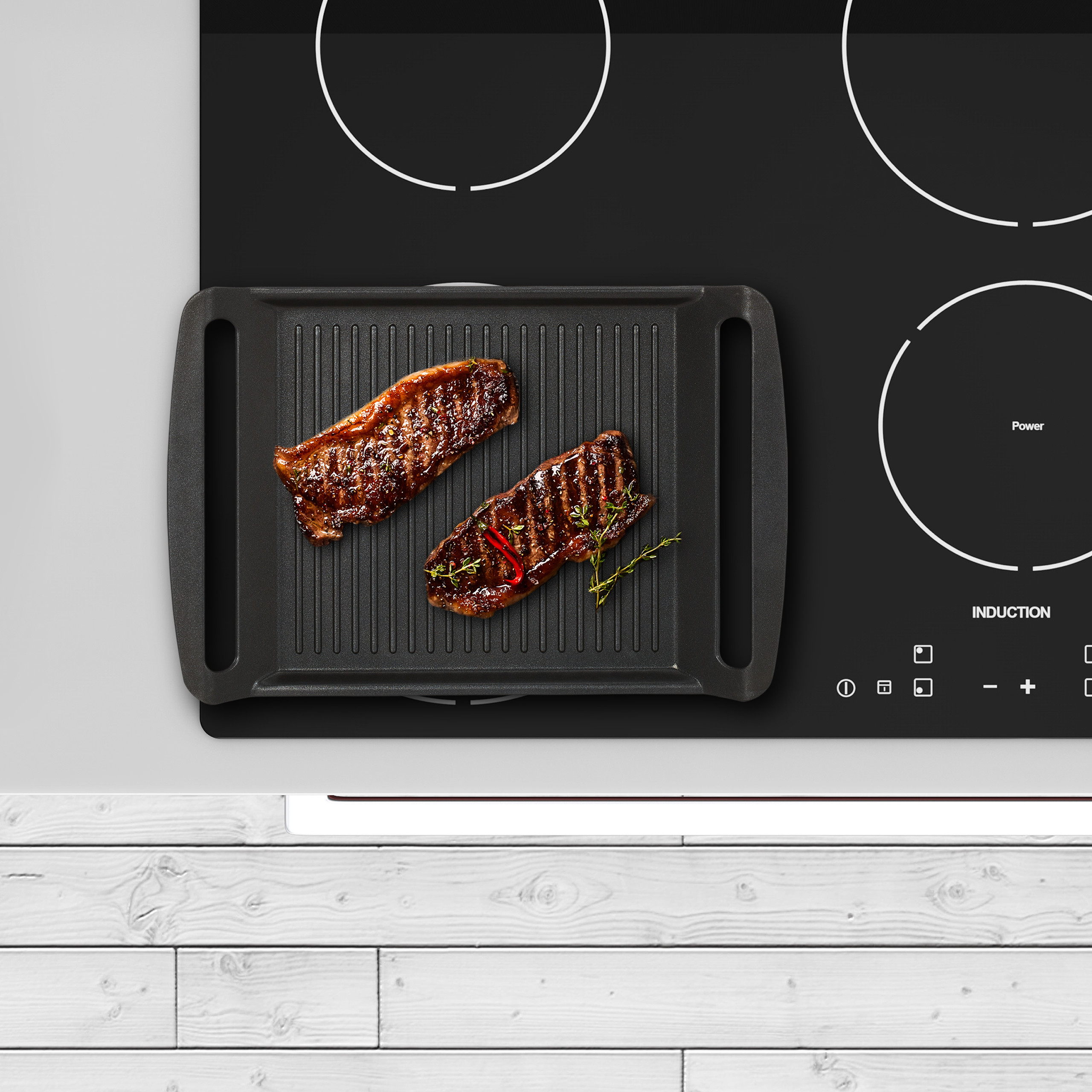 Zwei gebratene Steaks liegen in einer schwarzen Grillpfanne auf einem Induktionskochfeld mit weißem Holzuntergrund.