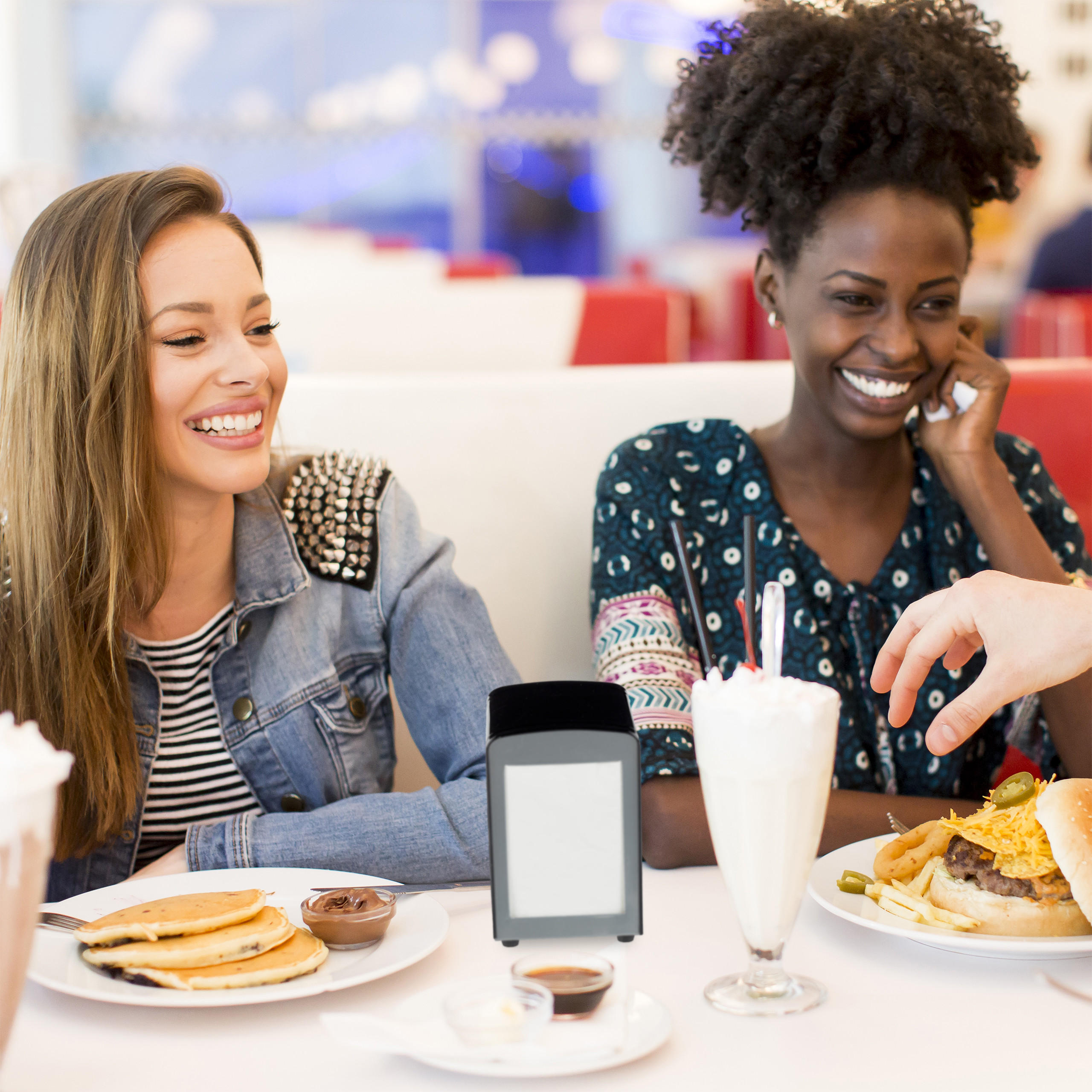 Zwei lächelnde junge Frauen sitzen in einem Imbiss an einem Tisch mit Pfannkuchen, einem Burger und Milchshakes.