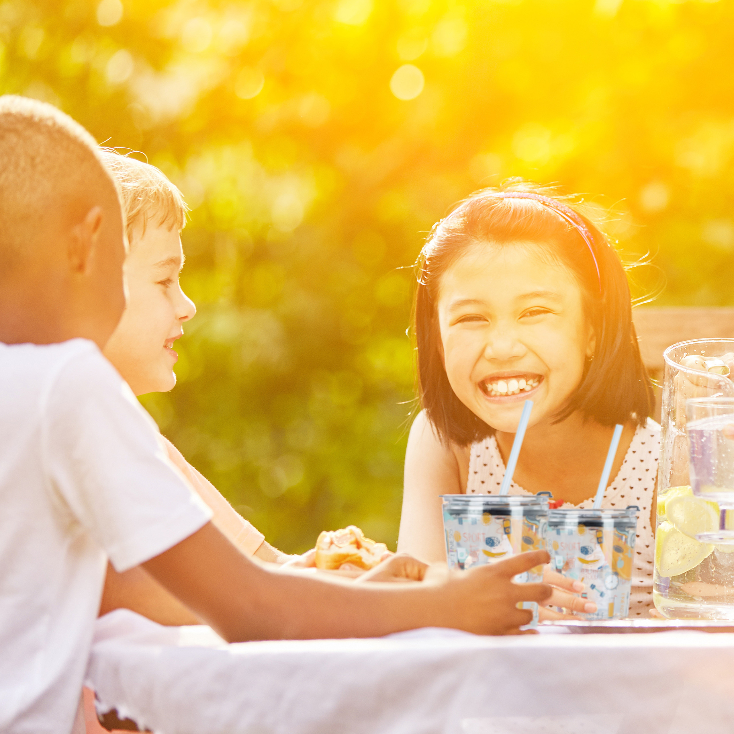 Kinder lachen und trinken an einem sonnigen Tisch im Freien.