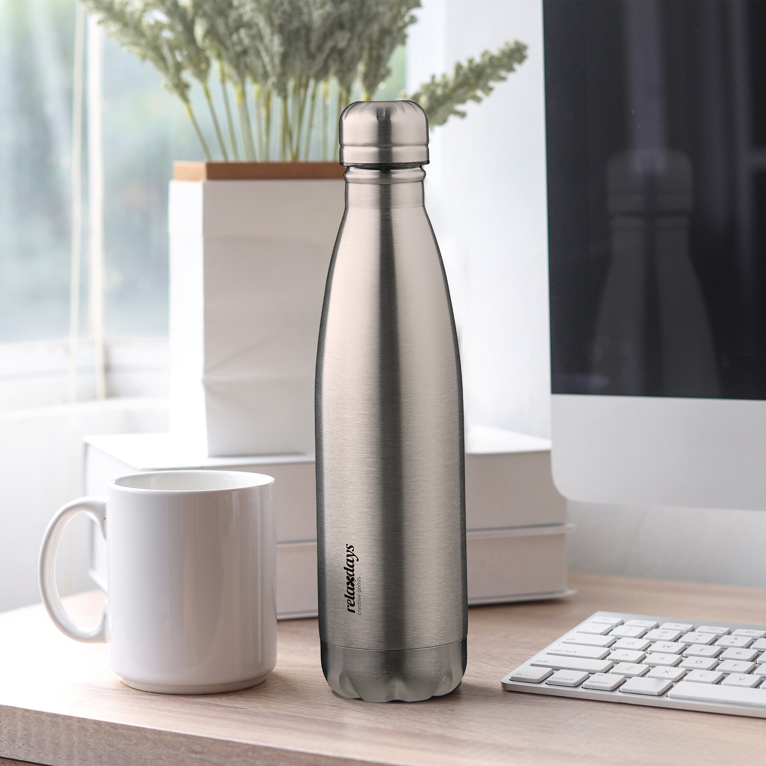 Eine silberne Thermosflasche auf einem Schreibtisch neben einer weißen Tasse, einem Computer und Pflanzen.