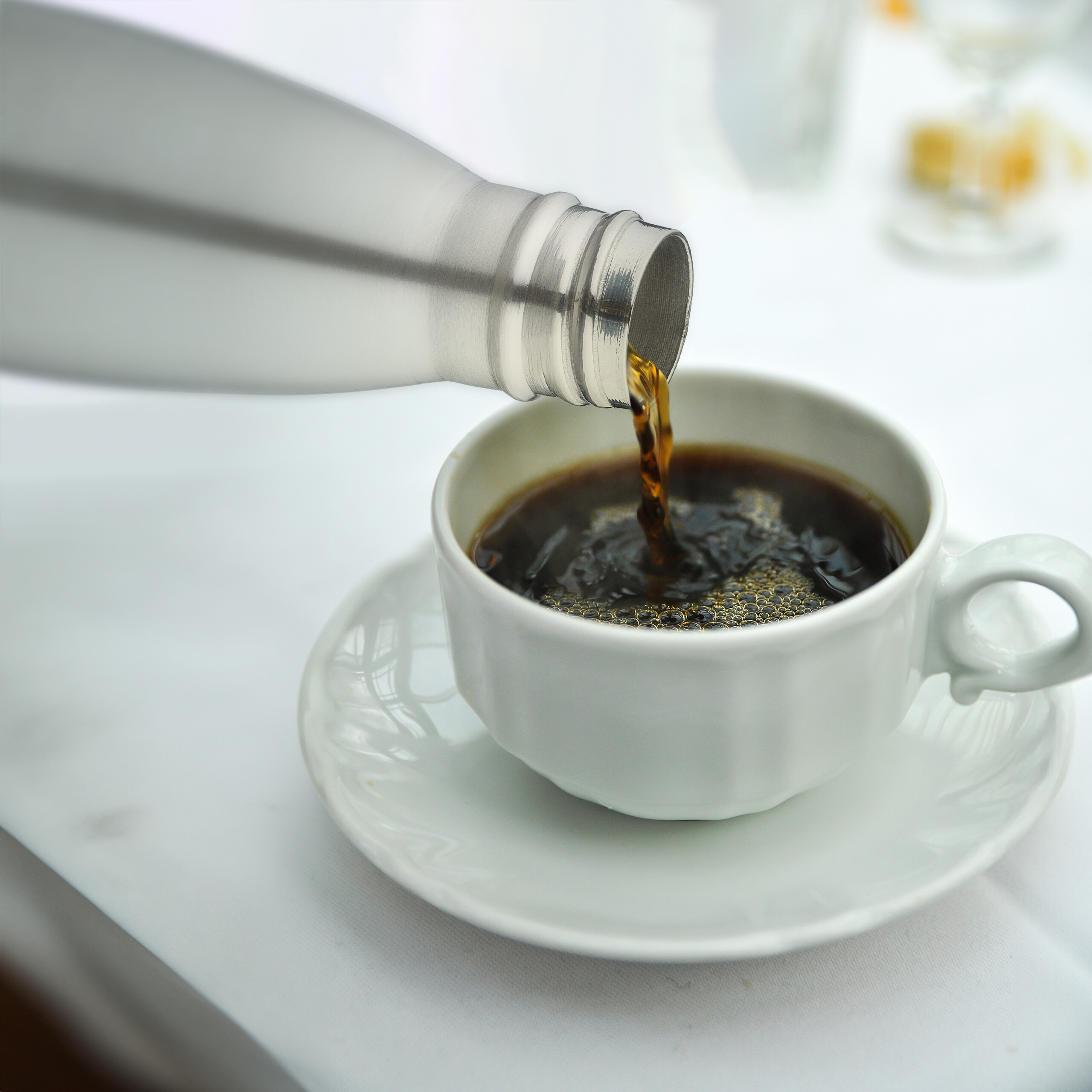 Eine Silberne Thermoskanne gießt Kaffee in eine weiße Tasse auf einer Untertasse.