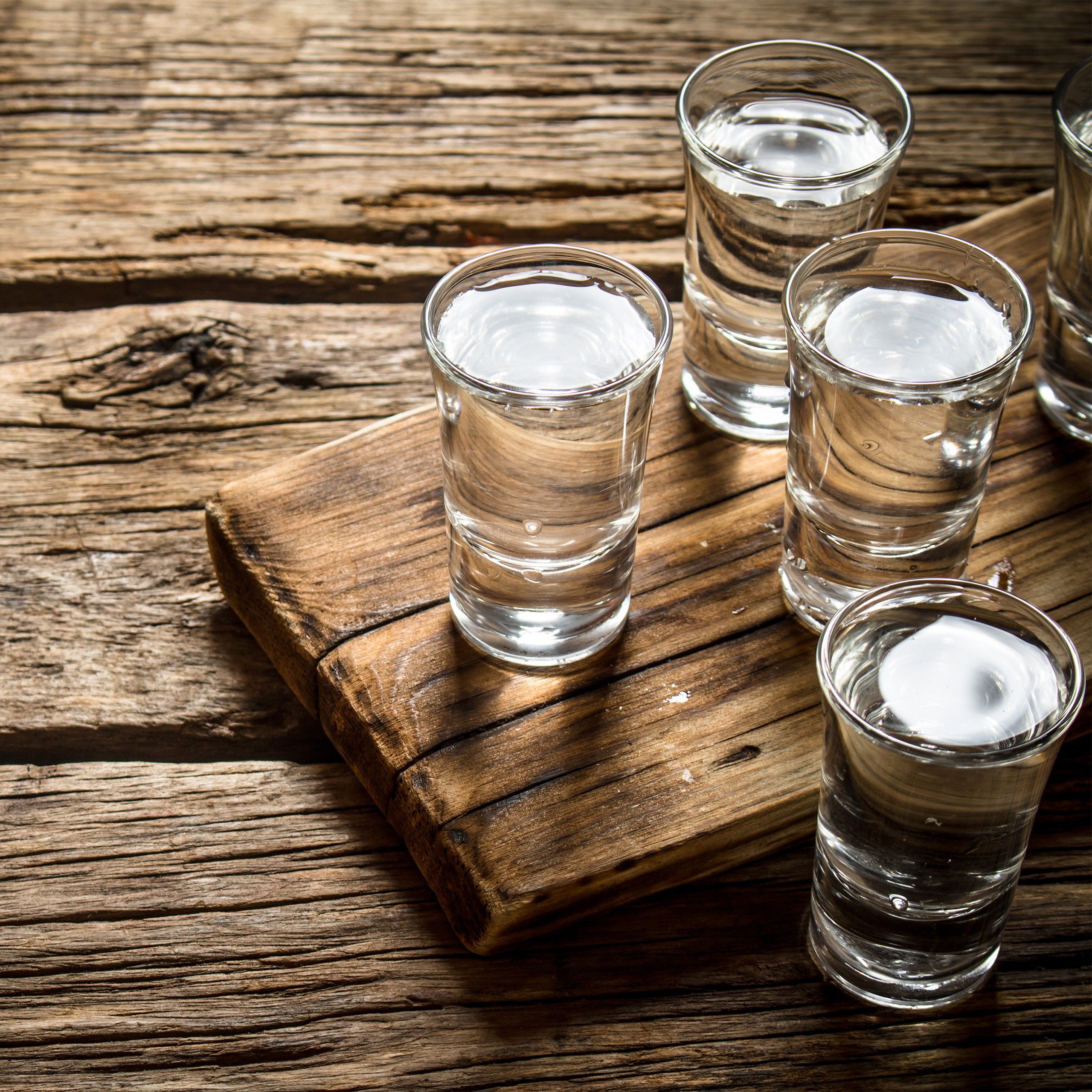 Vier Gläser klarer Schnaps auf einem hölzernen Servierbrett auf einem rustikalen Holztisch.