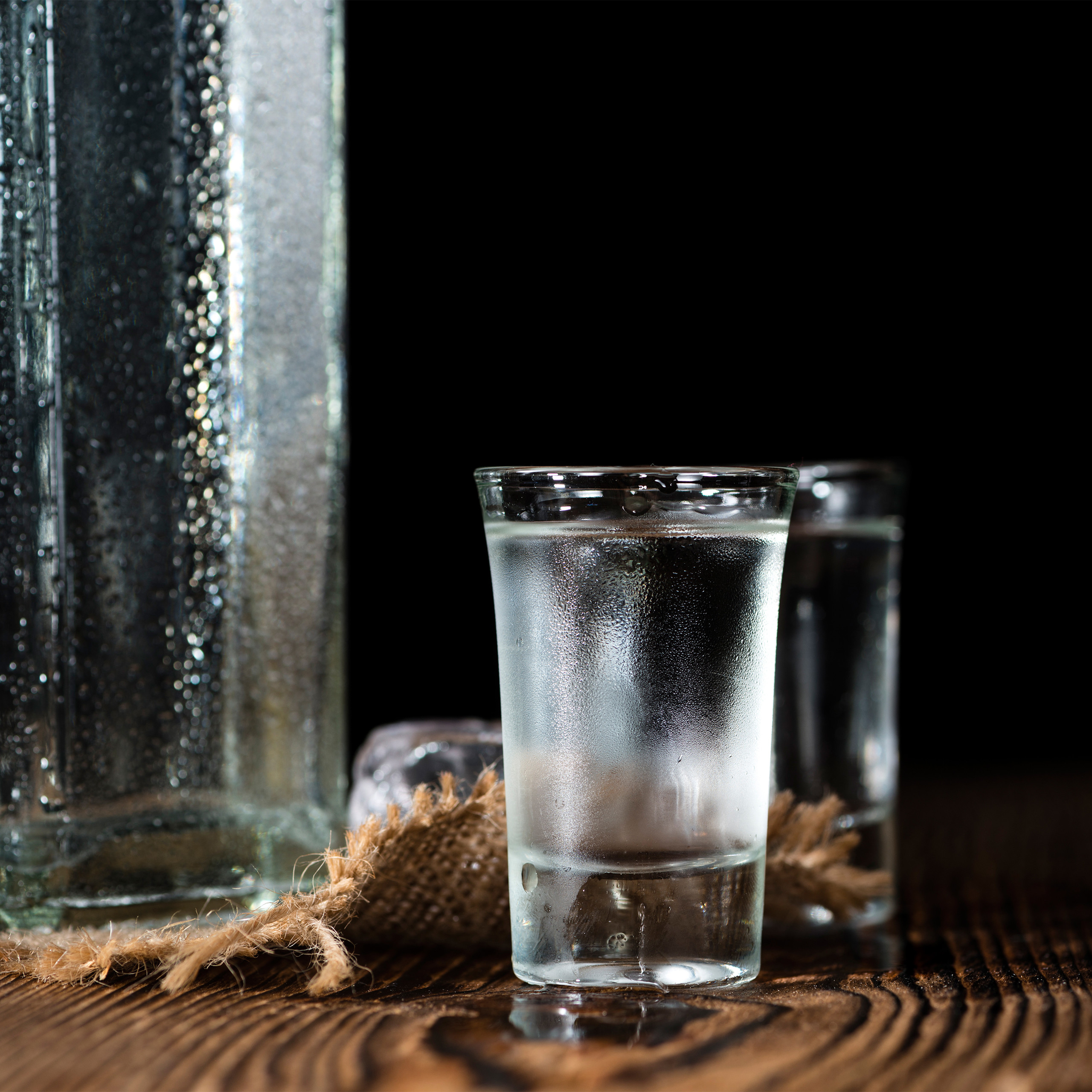 Ein kühles Schnapsglas mit klarem Geist steht auf dunklem Holz, mit nasser Flasche und Jute im Hintergrund.