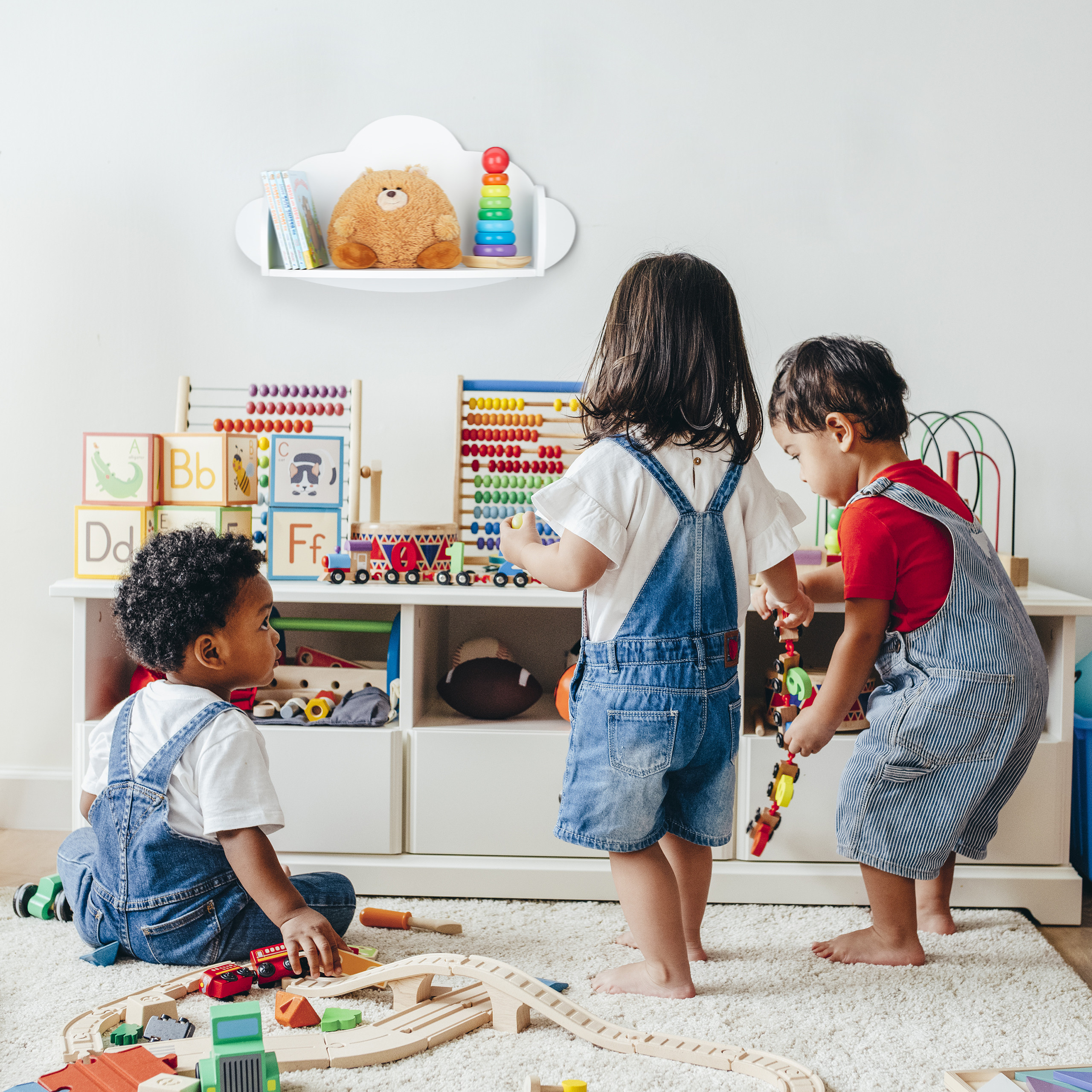 Drei Kleinkinder spielen in einem hellen Zimmer mit Spielzeug, während an der Wand ein Wolkenregal hängt.