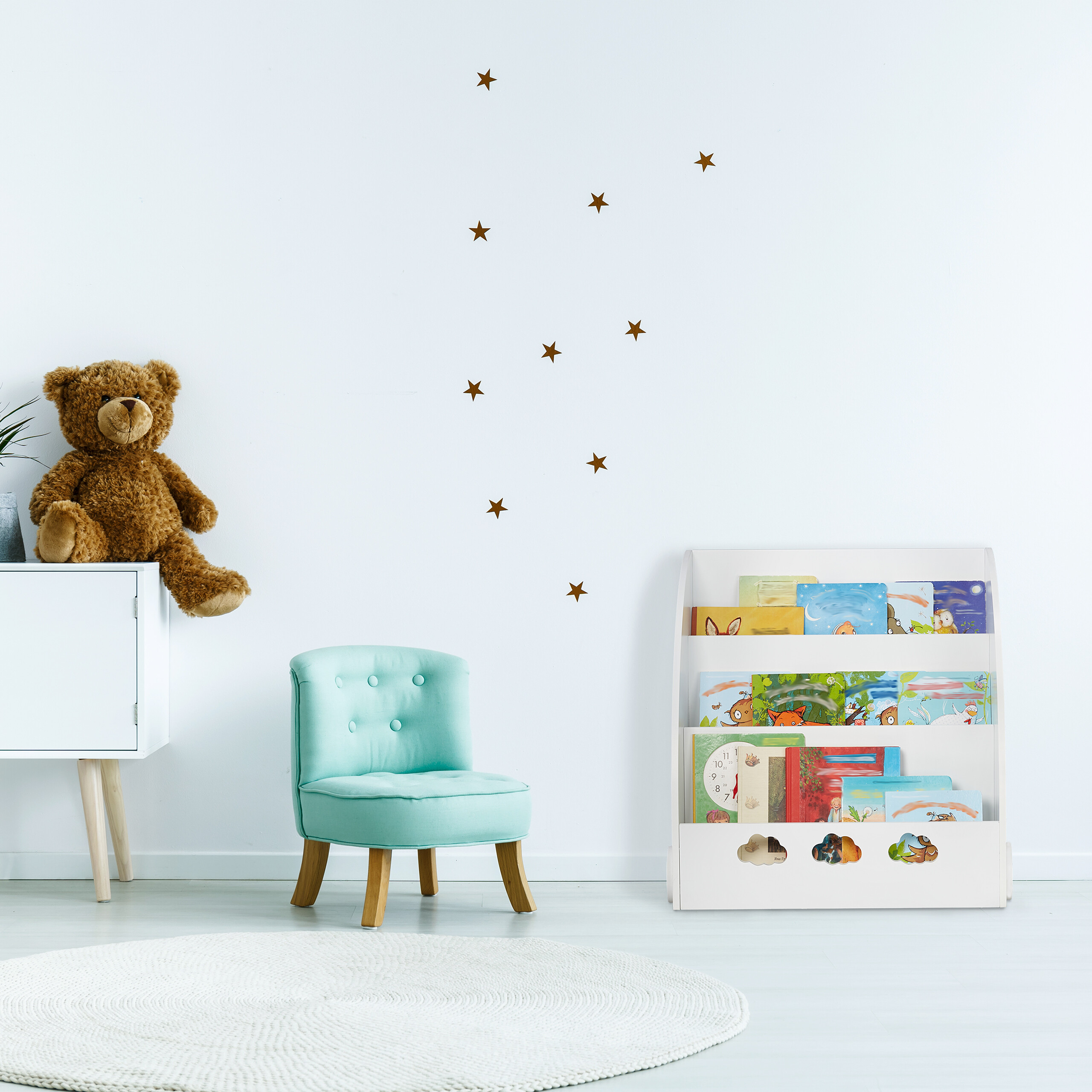 Ein Kinderzimmer mit einem Teddybären auf einem weißen Schrank, einem blauen Sessel und einem vollen Bücherregal.