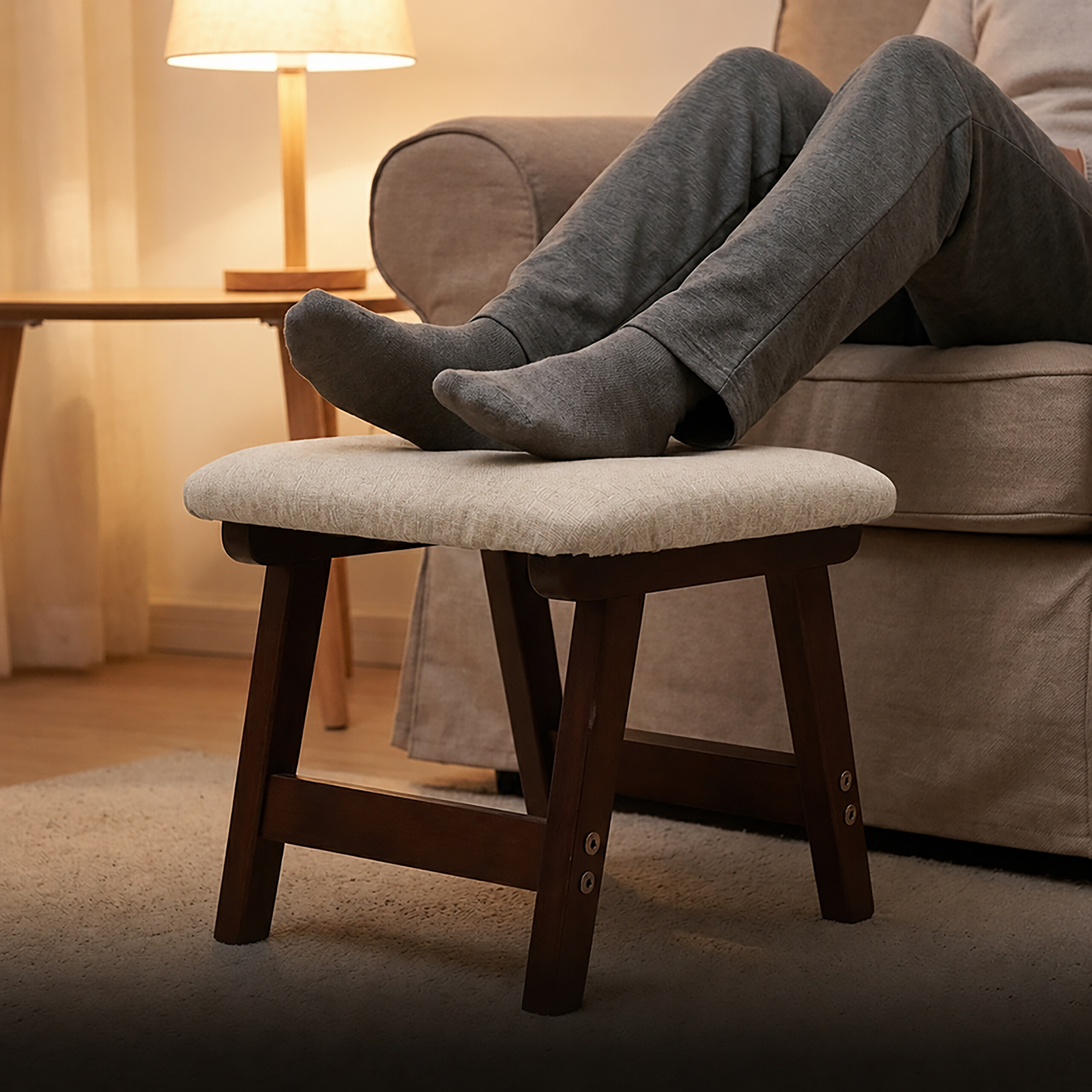 Eine Person in grauen Jogginghosen und Socken ruht die Füße auf einem kleinen Holzhocker in einem Wohnzimmer.
