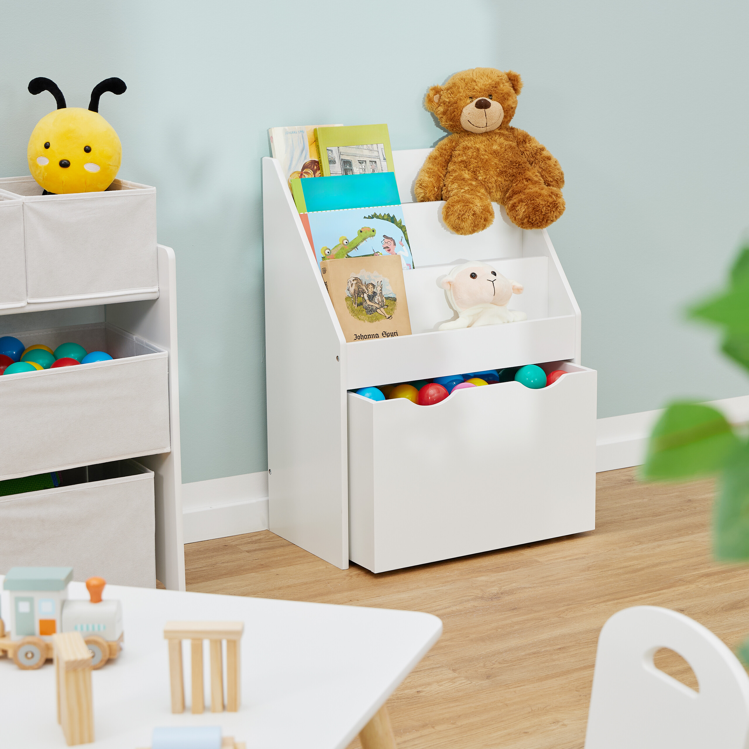 Ein weißes Kinderbücheregal mit Büchern, einem Teddybären, einem Plüschschaf und bunten Bällen in einem Spielzimmer.