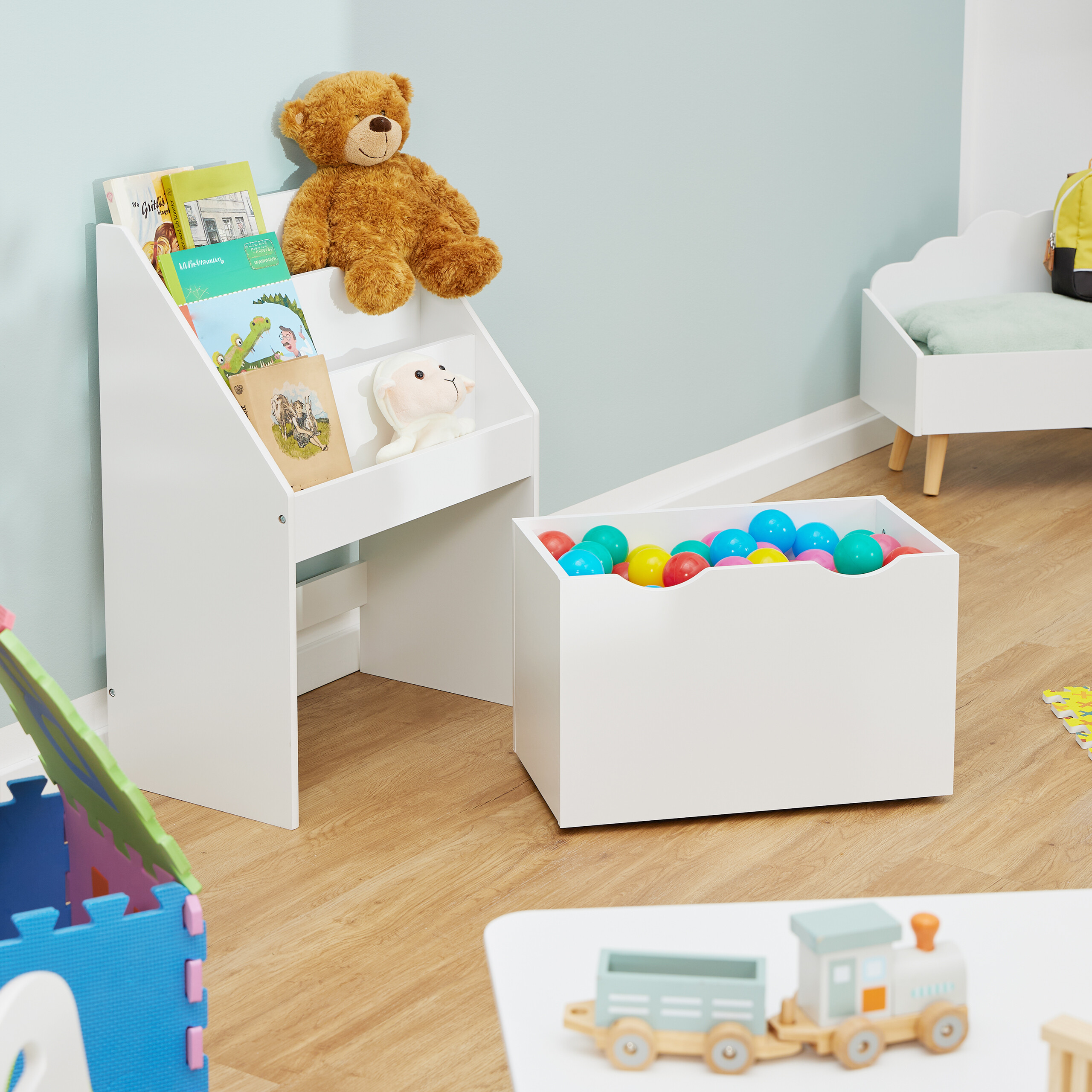 Kinderzimmer mit einem weißen Bücherregal voller Bücher und Kuscheltiere, einer Spielzeugkiste mit bunten Bällen und Holzspielzeug.