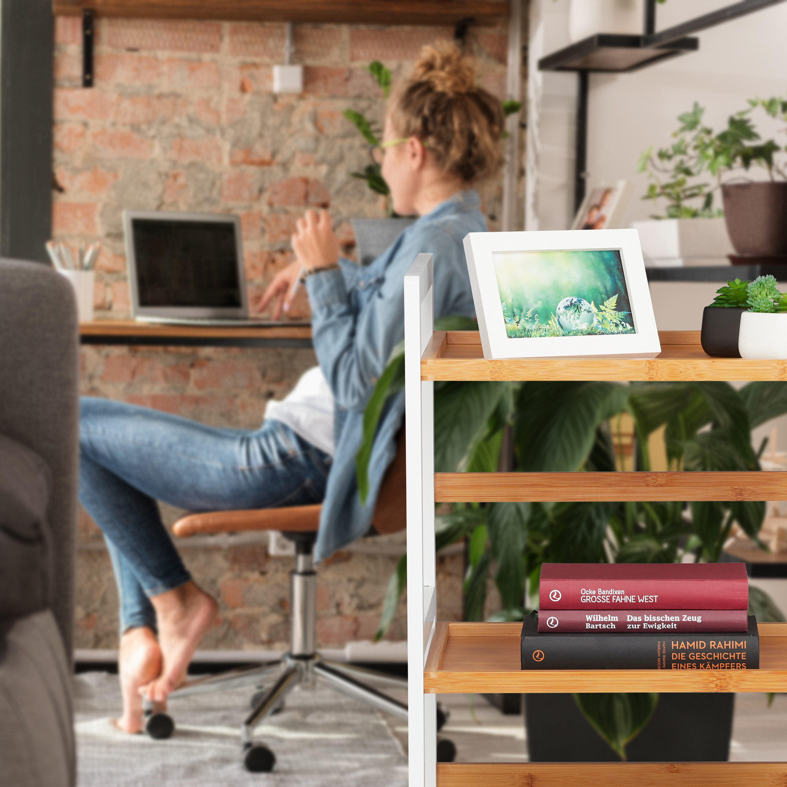 Ein hölzernes Regal mit Bild und Büchern im Vordergrund; im Hintergrund arbeitet eine Frau am Laptop vor einer Ziegelwand.