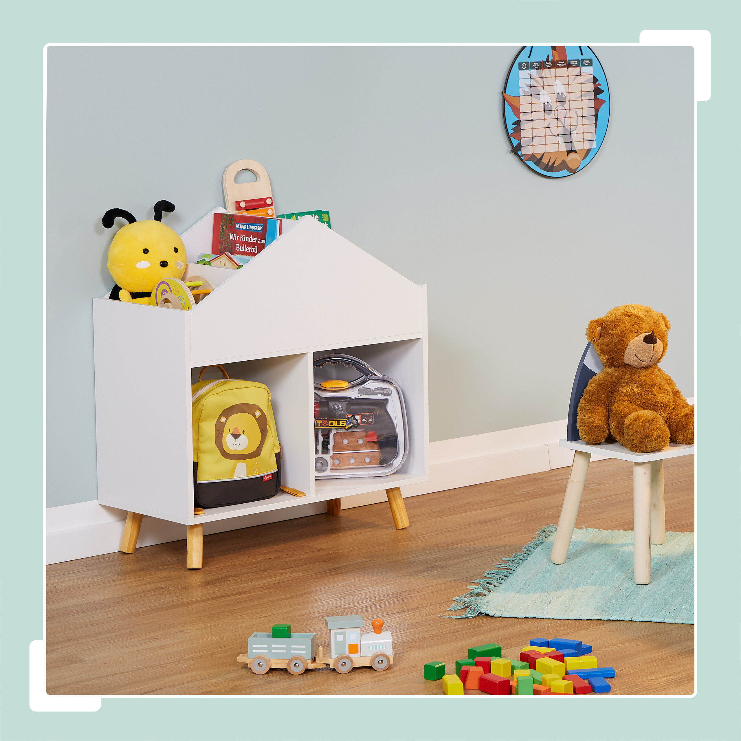 Ein Kinderzimmer mit weißem Hausregal, Teddybär auf Stuhl, Holzeisenbahn und Bauklötzen auf dem Boden.