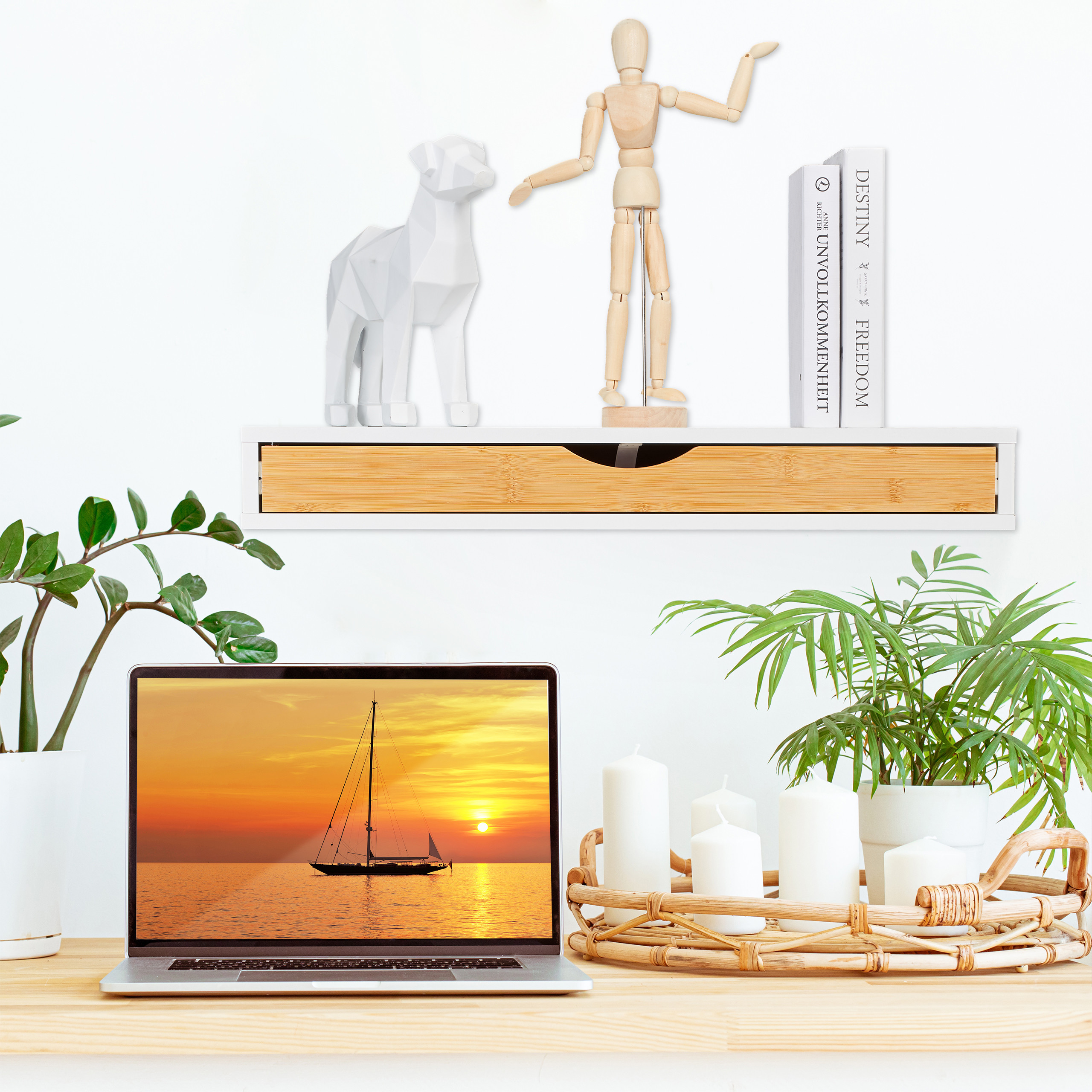 Schreibtisch mit Laptop, Pflanzen, Kerzen auf Tablett und einem Regal mit Deko an der weißen Wand.