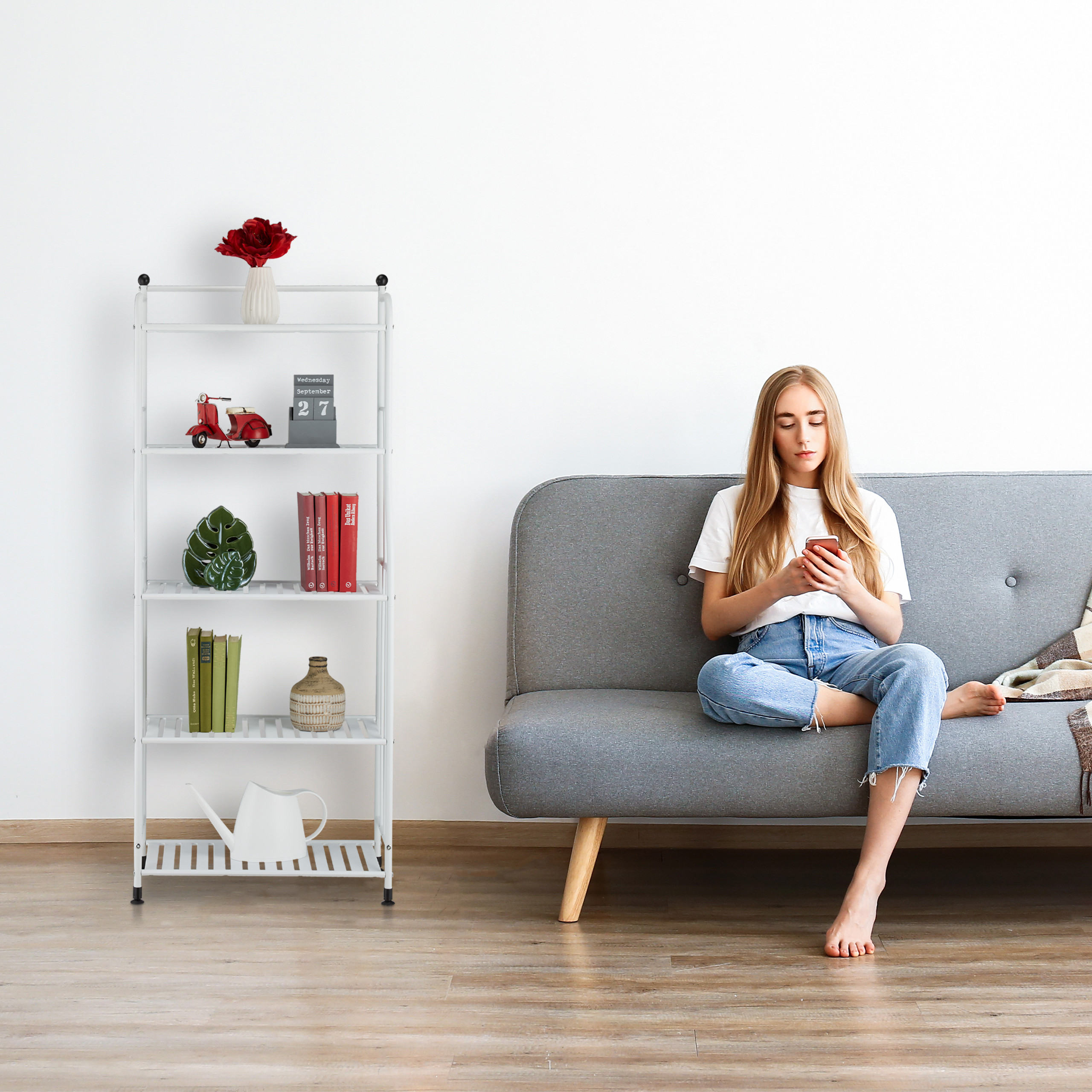 Eine blonde Frau sitzt auf einem grauen Sofa und nutzt ihr Smartphone. Neben ihr steht ein weißes Regal mit Deko.