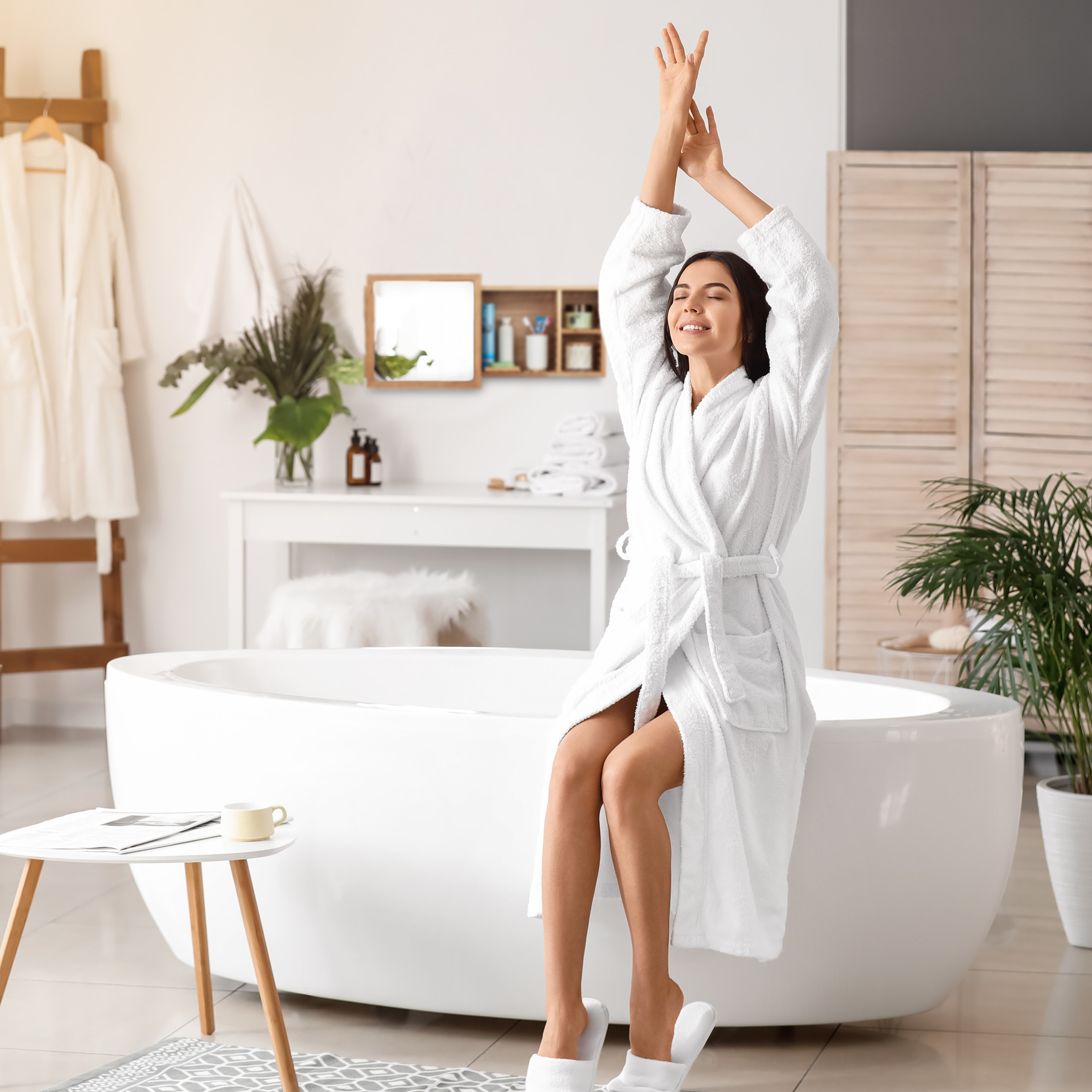 Eine Frau im weißen Bademantel sitzt auf einer Badewanne und reckt die Arme in einem hellen Badezimmer nach oben.