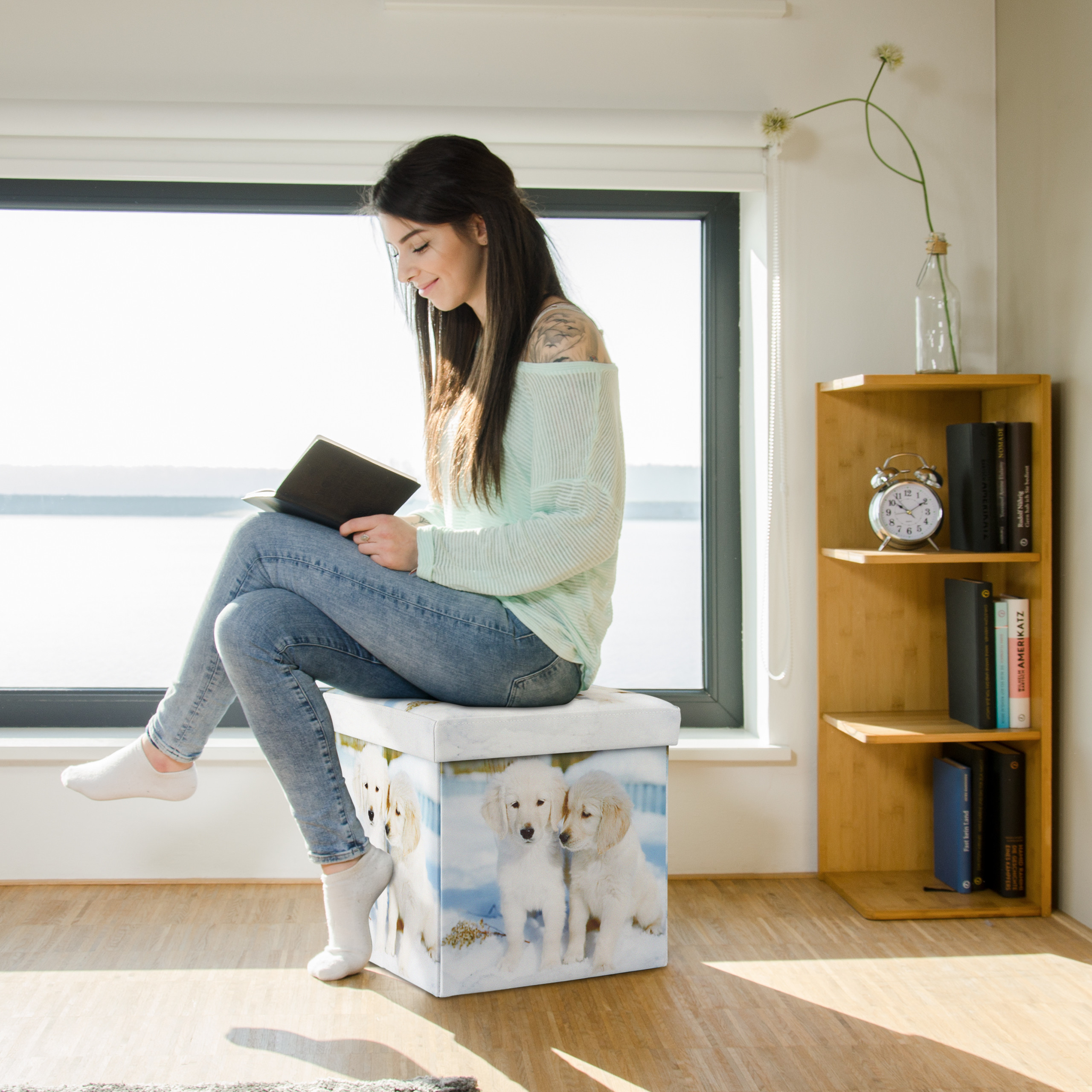 Eine Frau sitzt auf einem bedruckten Hocker vor einem Fenster und liest ein Buch.