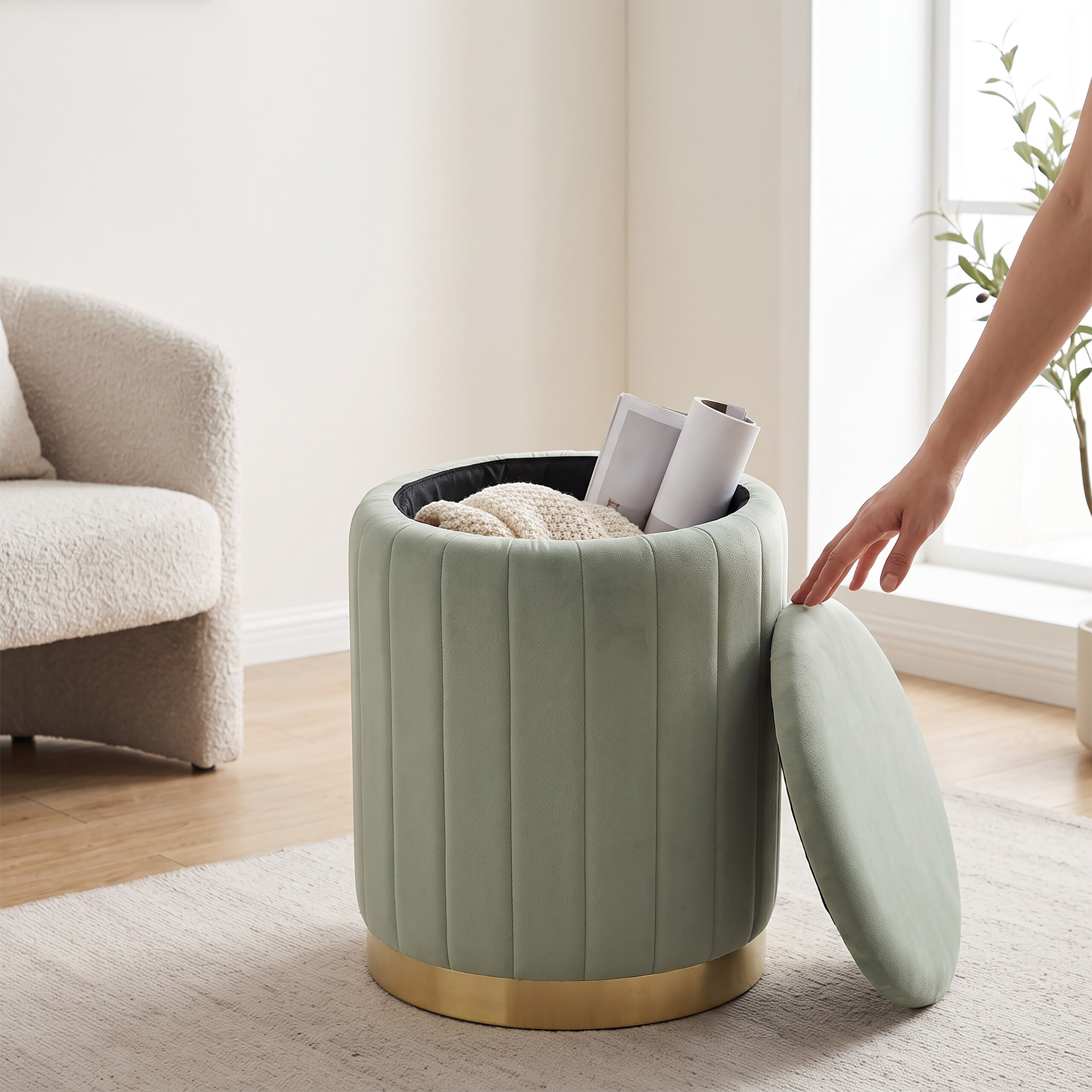 Eine Hand öffnet einen grünen Stauraum-Hocker mit Goldbasis, der Decke und Zeitschriften enthält.