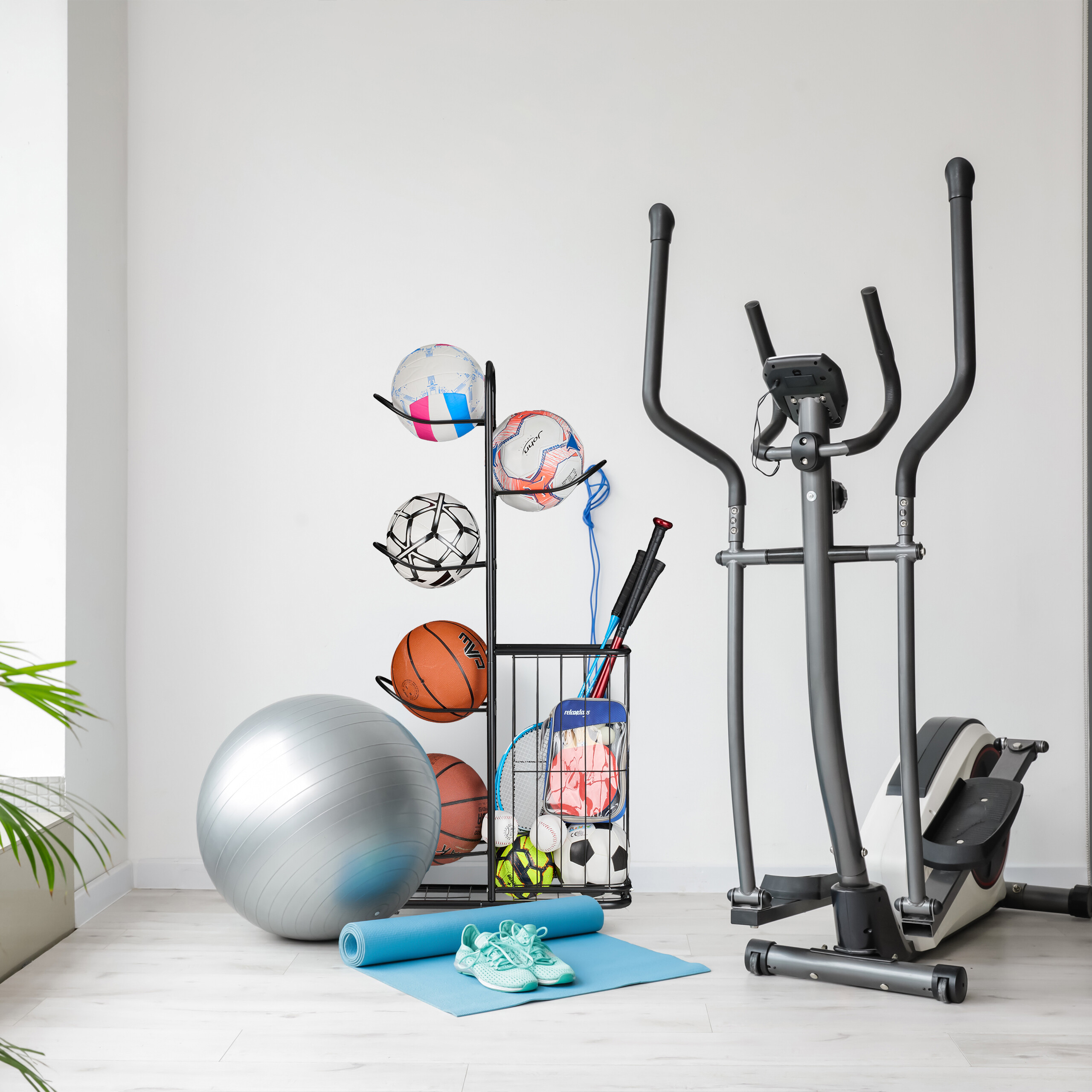 Ein Fitnessstudio mit Ellipsentrainer, Bällen, Yogamatte und Laufschuhen vor einer weißen Wand.