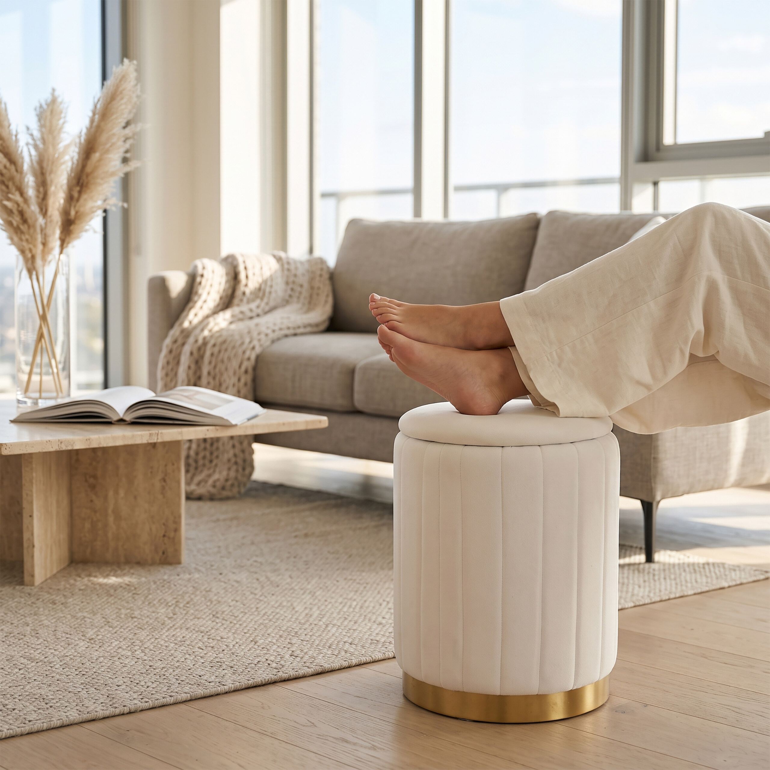 Barfüße ruhen auf einem weißen Samthocker in einem hellen Wohnzimmer mit Sofa und großen Fenstern.