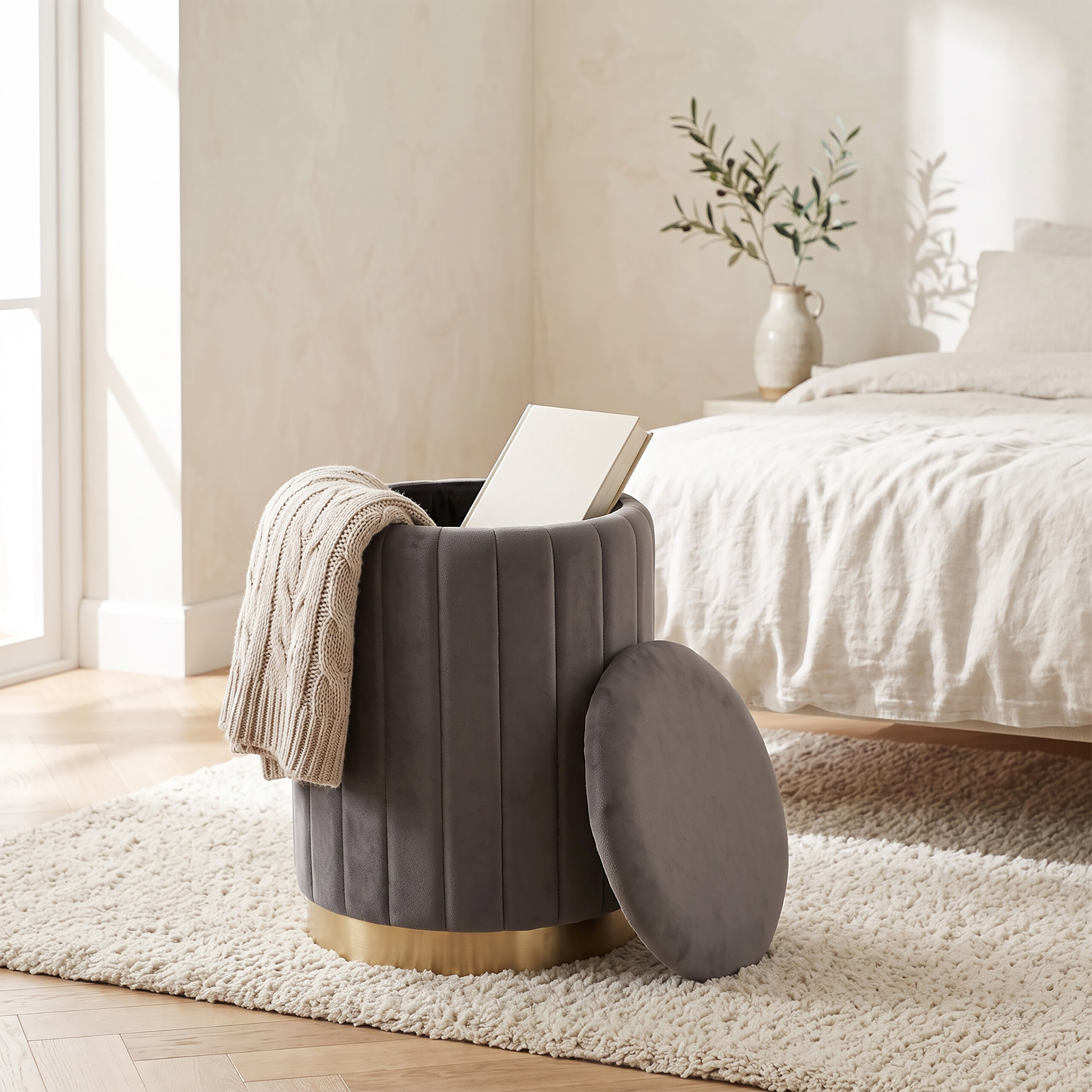 Grauer samtartiger Hocker mit goldener Basis, Buch und Decke im hellen Schlafzimmer.