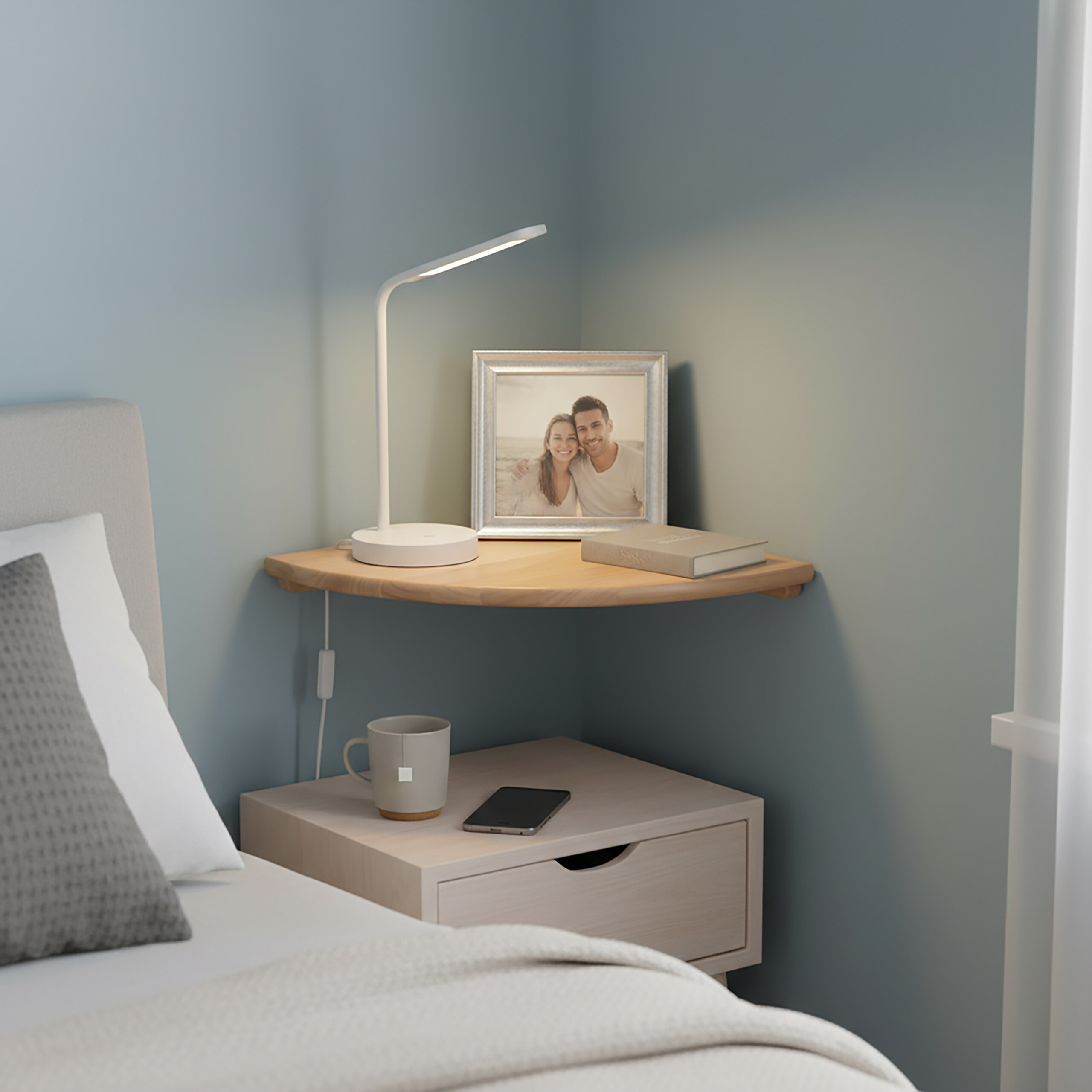 Eckregal mit Lampe, Bilderrahmen und Buch über Nachttisch, Tasse und Smartphone in Schlafzimmer.