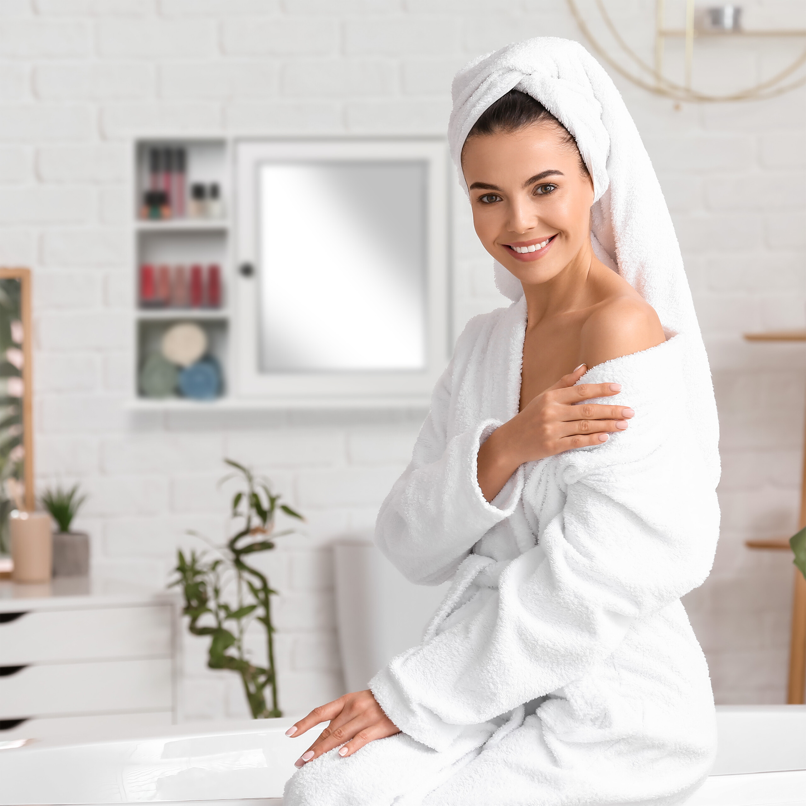 Eine lächelnde Frau im weißen Bademantel und Handtuch sitzt in einem hellen Badezimmer.