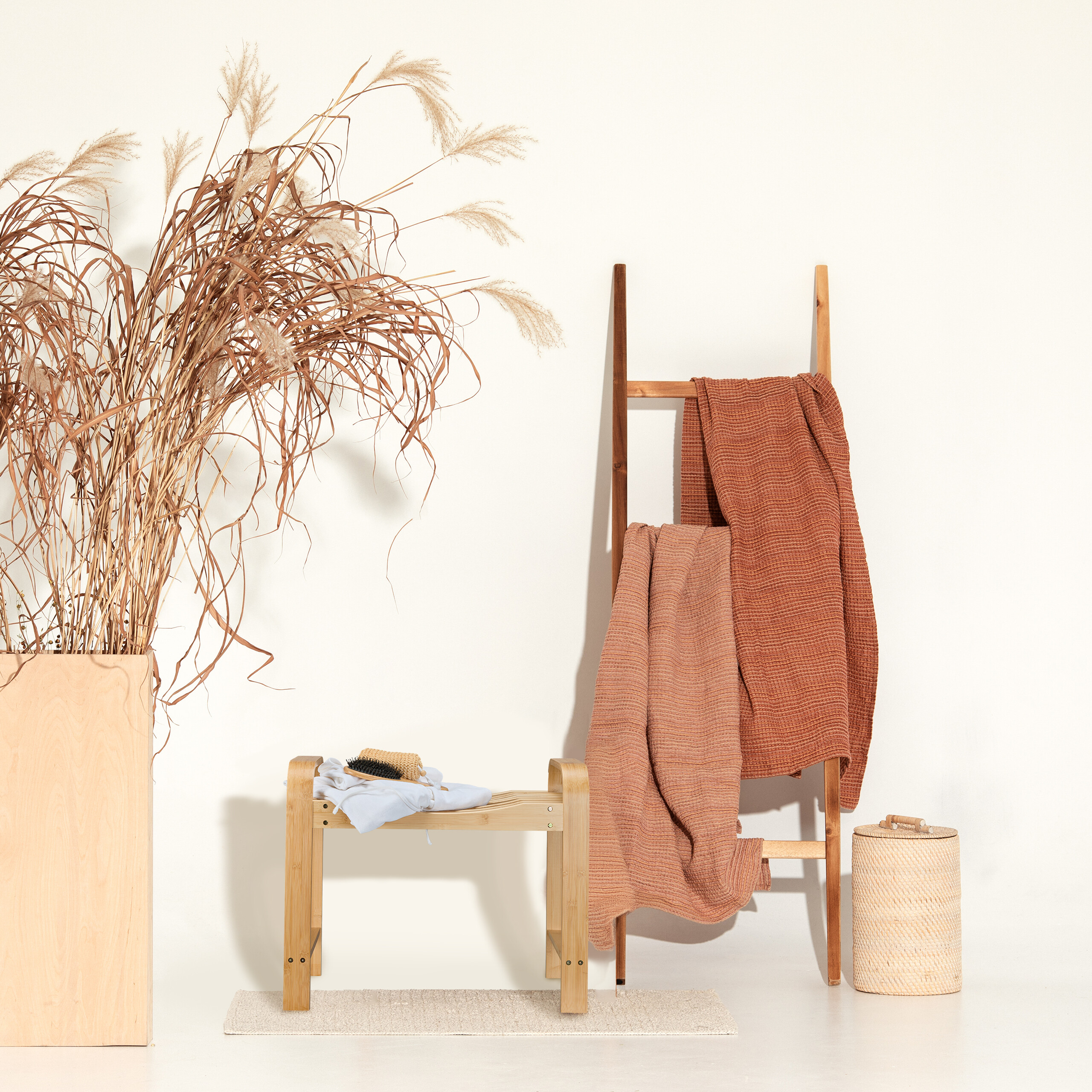 Holzleiter mit Handtüchern, Bambusbank mit Kleidung und Bürste, großer Pflanzkübel und Korb vor weißer Wand.