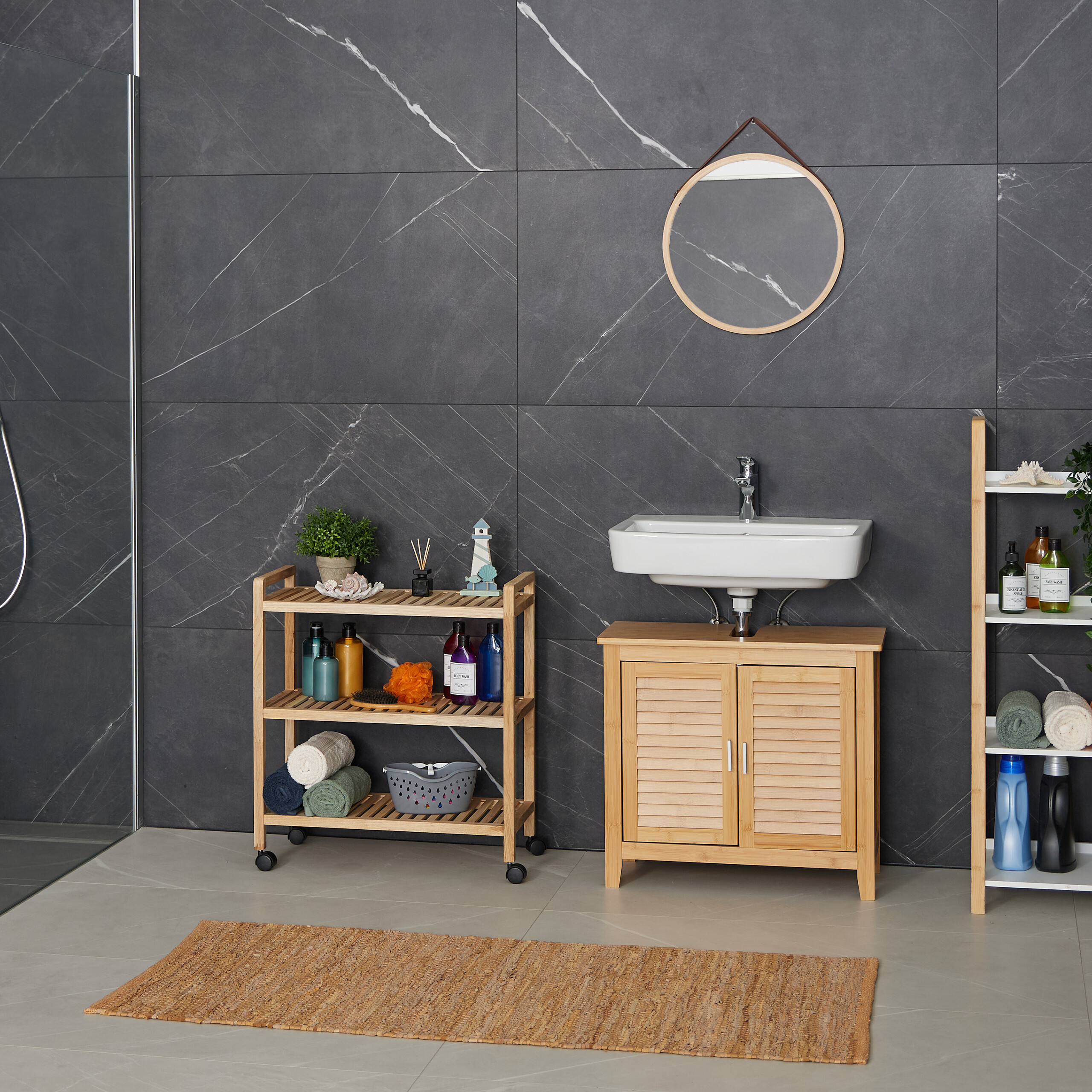 Ein modernes Badezimmer hat ein weißes Waschbecken, Bambusmöbel, einen runden Spiegel und dunkle Marmorfliesen.