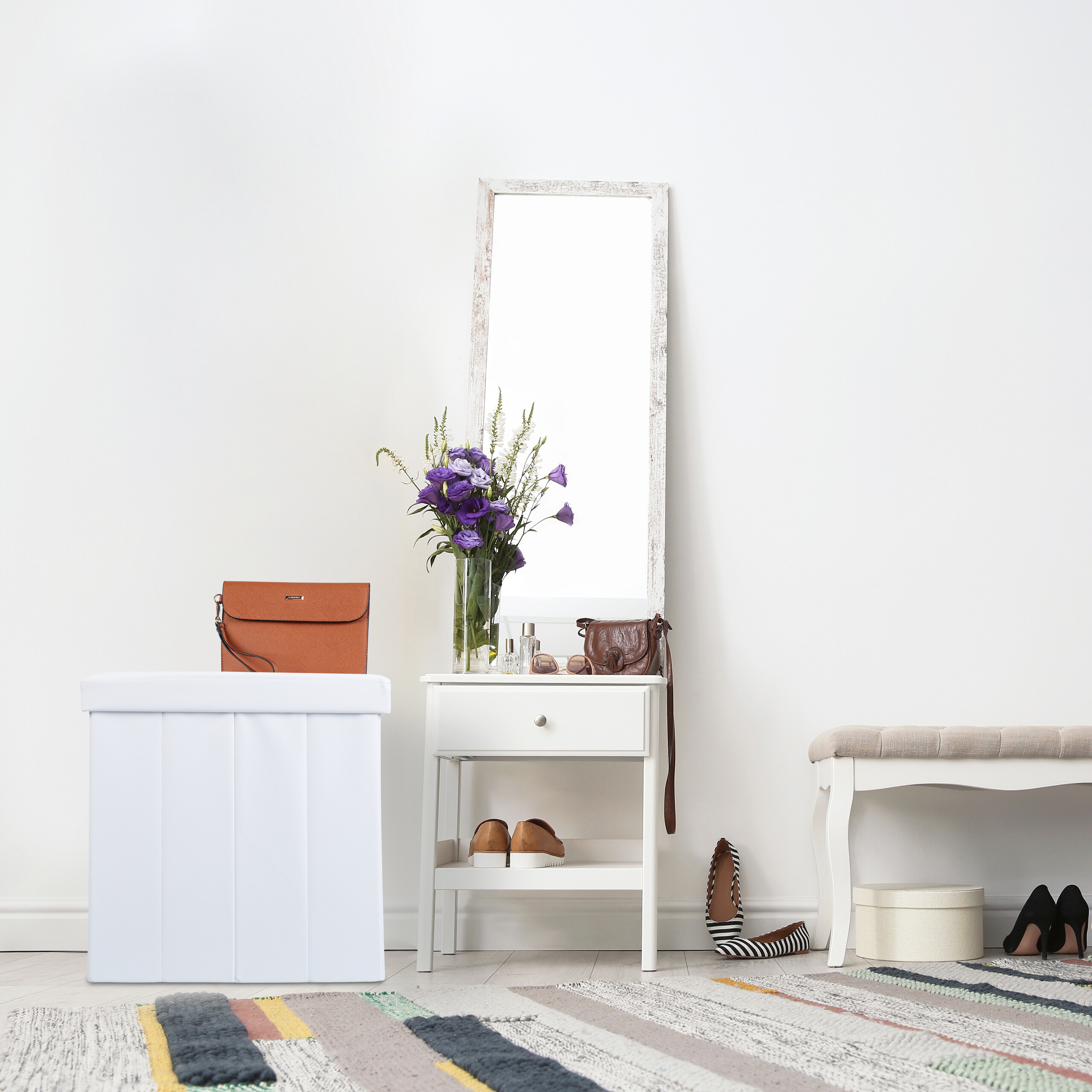 Ein heller Flur mit weißem Hocker, Konsole mit Spiegel, Blumen und Taschen sowie einer Bank auf gemustertem Teppich.