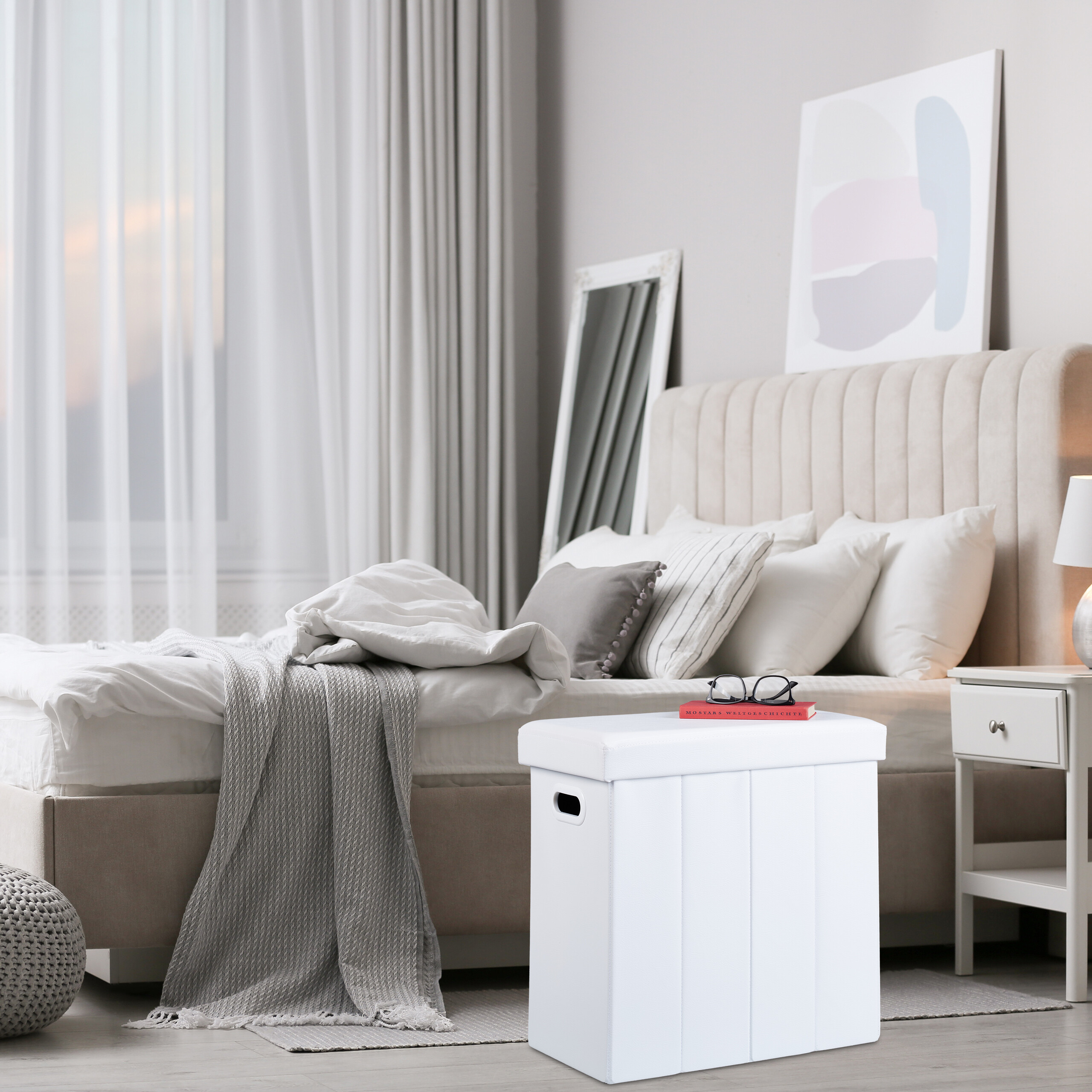 Ein weißer Hocker steht im Schlafzimmer vor einem beige gepolsterten Bett mit Kissen.