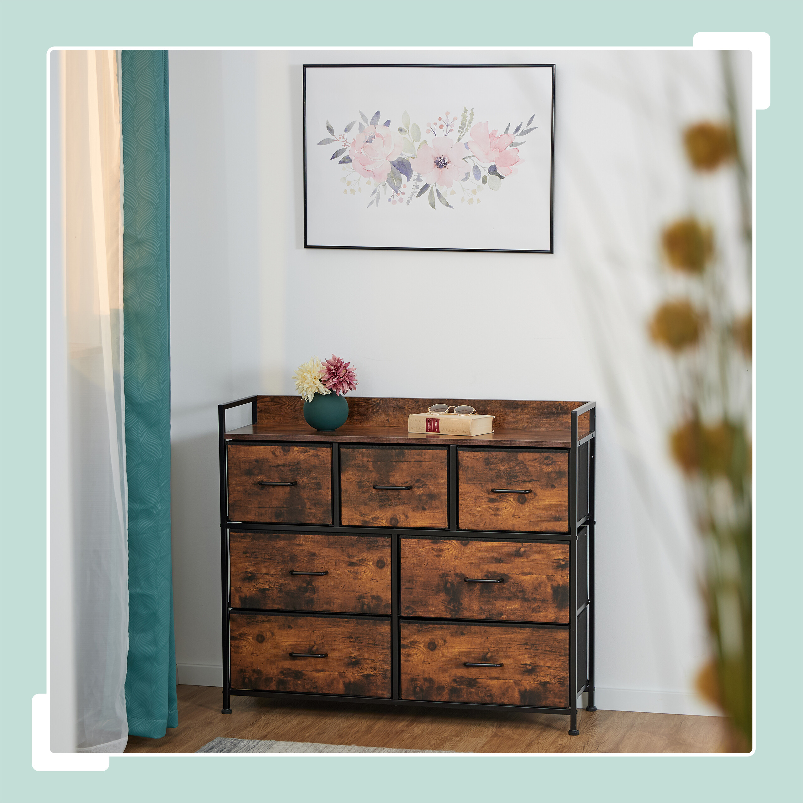 Eine hölzerne Kommode mit sieben Schubladen steht an einer weißen Wand, darüber hängt ein Blumenbild.