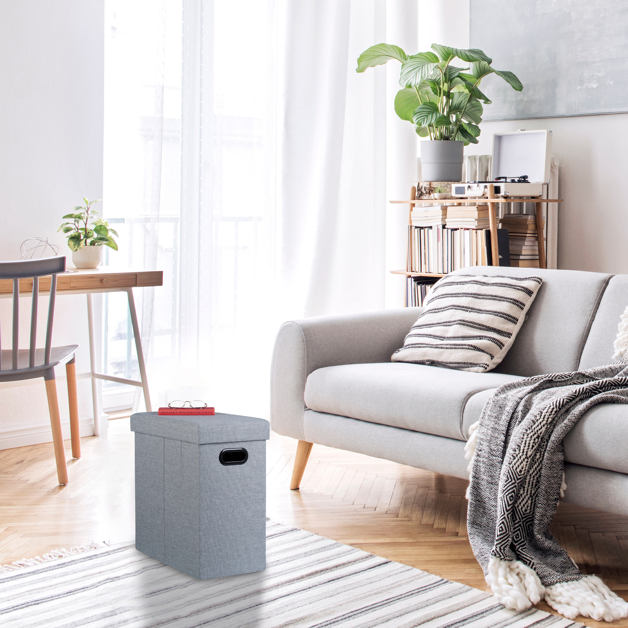 Ein grauer Stoffhocker mit Buch steht vor einem grauen Sofa in einem hellen Wohnzimmer mit Holzfussboden.