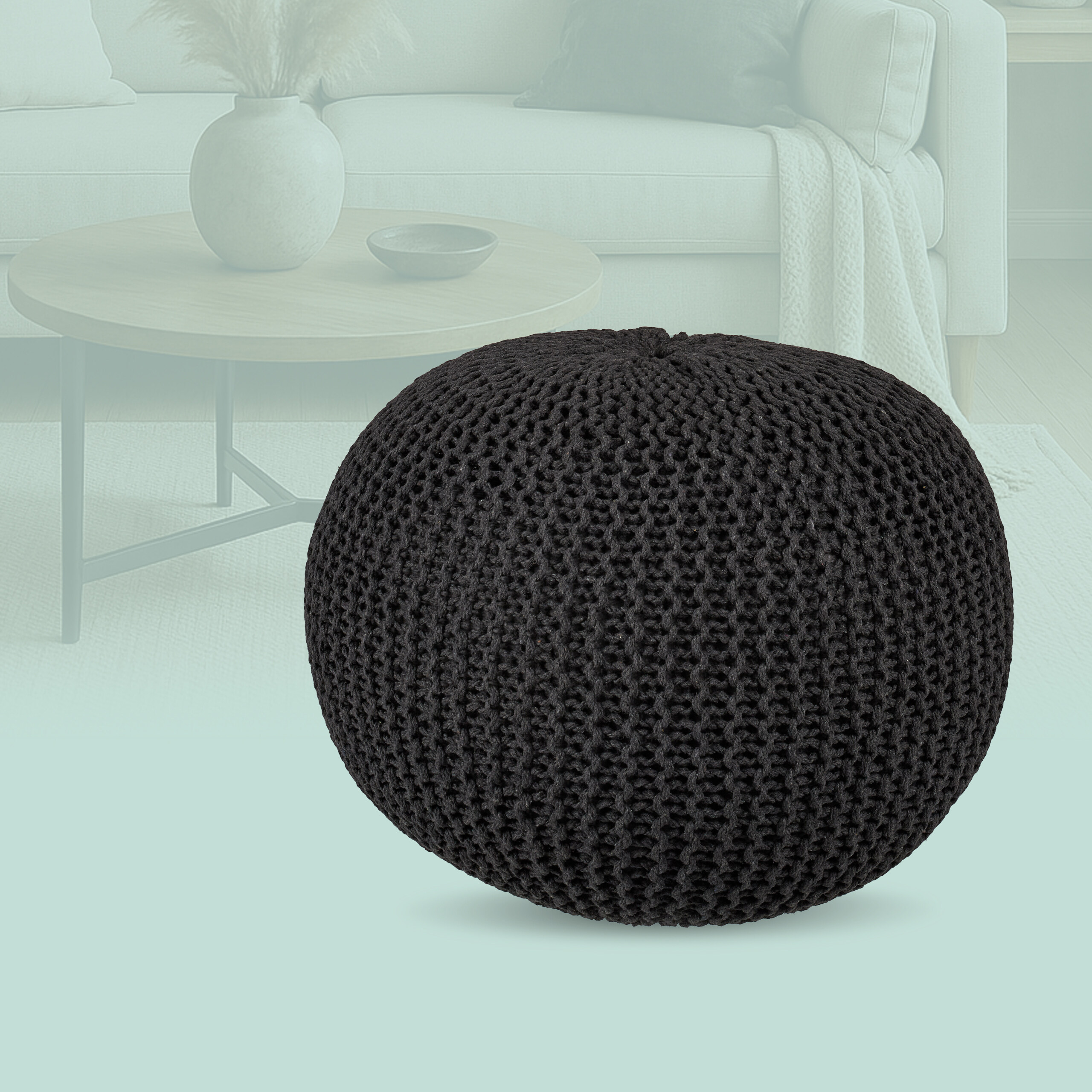 Ein runder, schwarzer Strickpouf steht im Vordergrund vor einem hellen Wohnzimmer mit Sofa und Tisch.