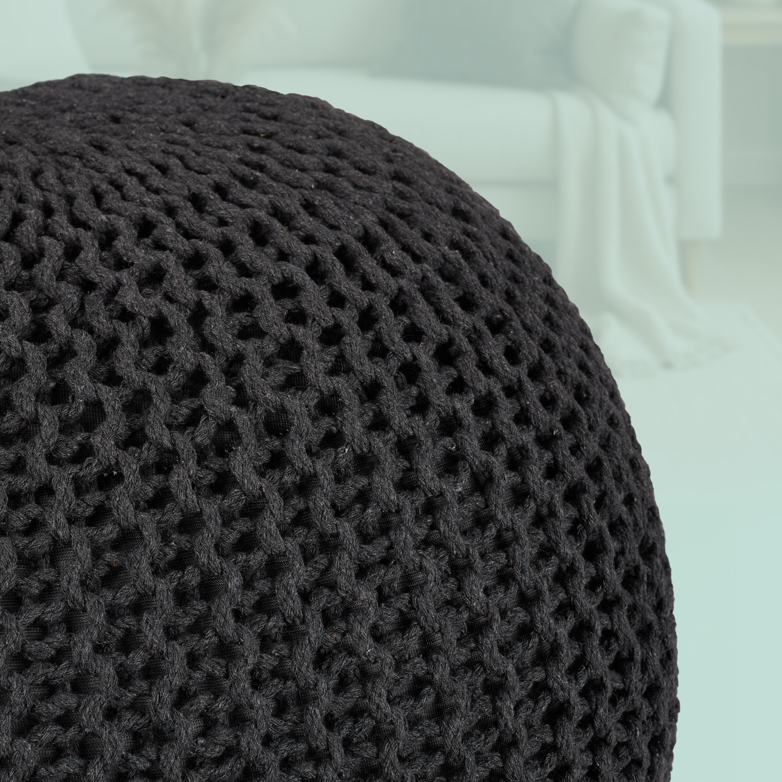Ein großer, dunkelgrauer gestrickter Pouf steht im Vordergrund, im Hintergrund ist ein helles Zimmer zu sehen.