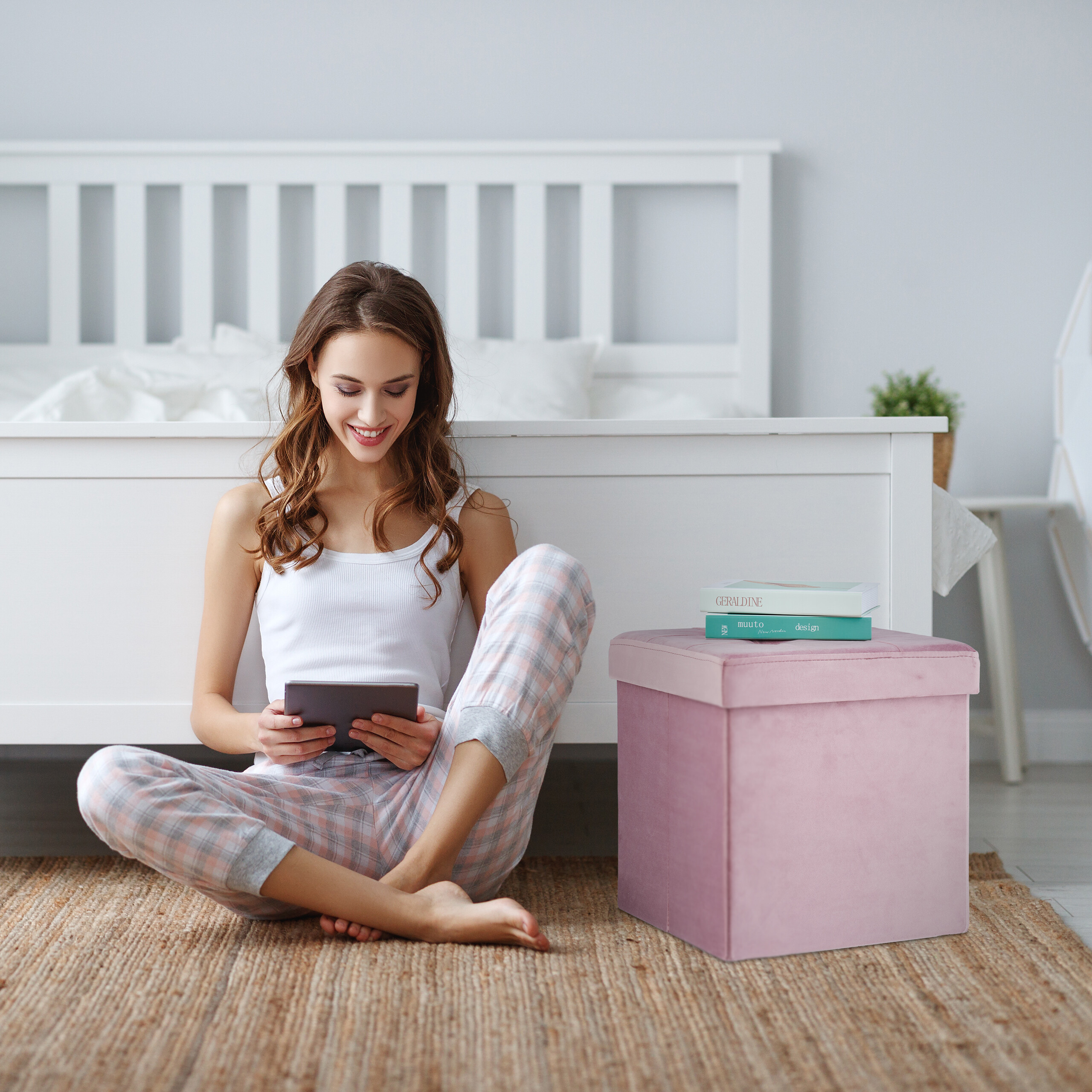 Eine Frau sitzt im Schlafzimmer auf dem Boden, nutzt ein Tablet, neben ihr ein rosa Hocker mit Büchern.