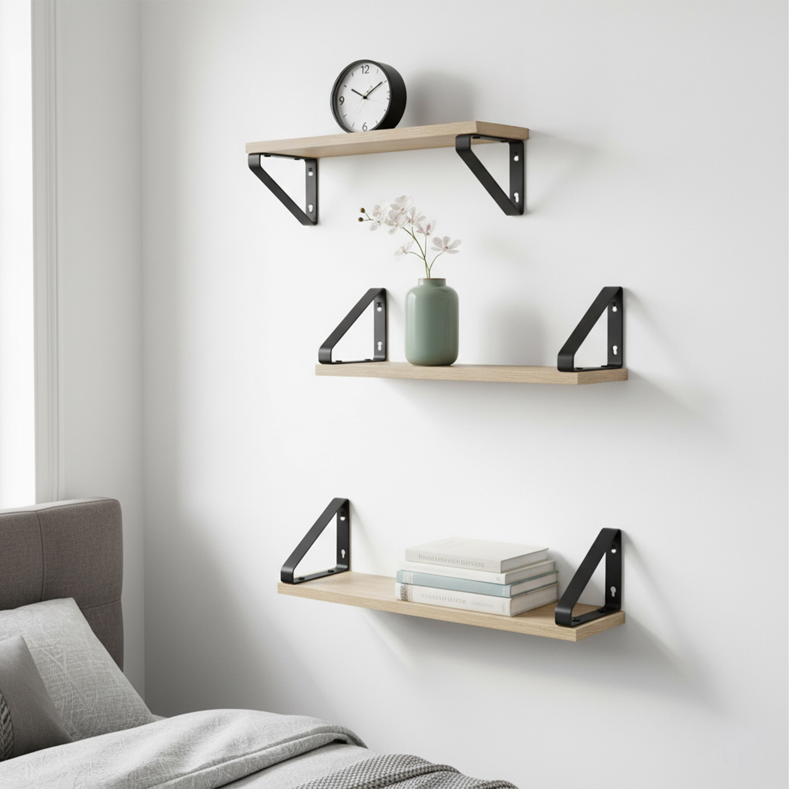 Drei Holzwandregale mit schwarzen Halterungen an einer weißen Wand über einem Bett. Deko: Uhr, Vase und Bücher.