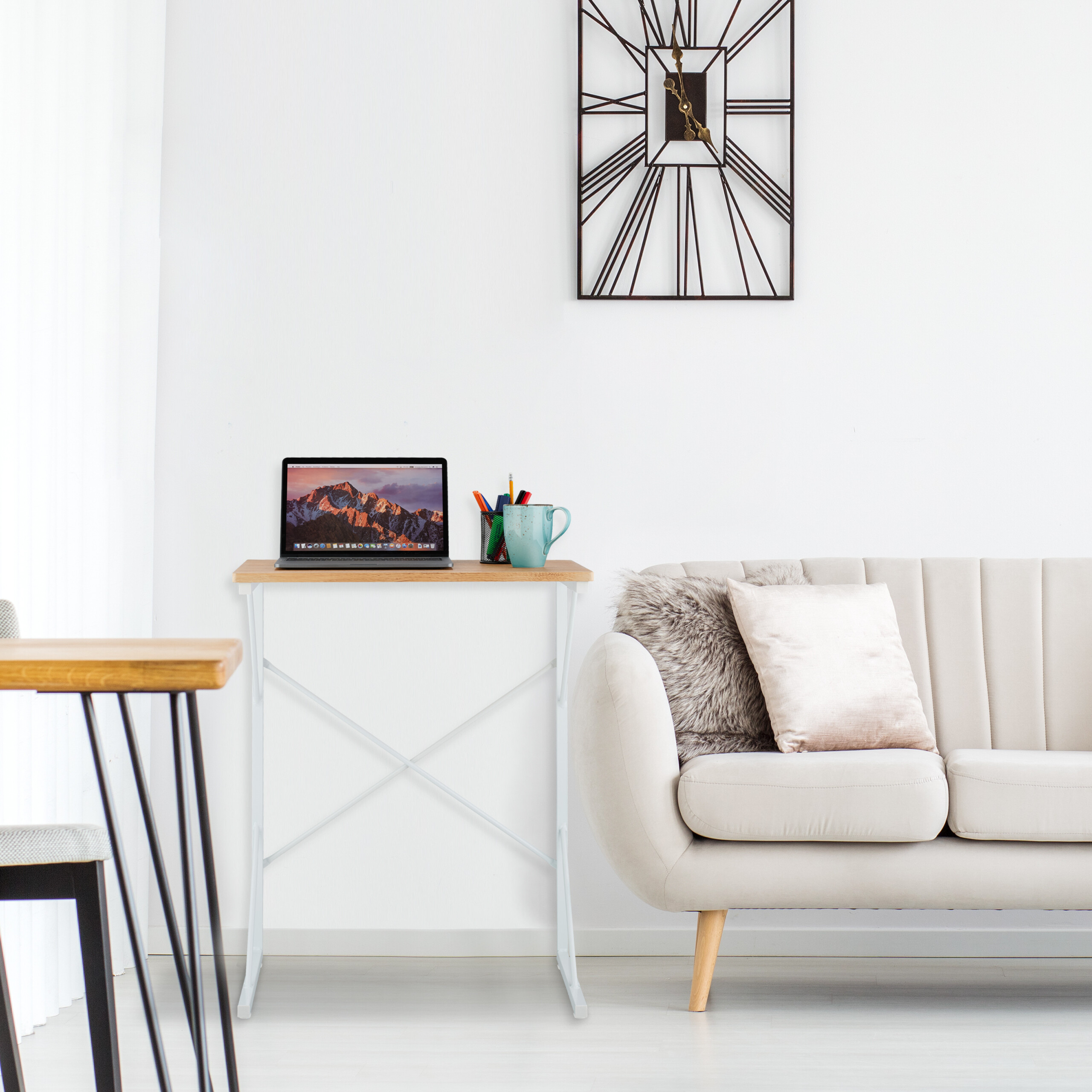 Laptop auf kleinem Schreibtisch neben beigem Sofa mit Kissen und moderner Wanduhr.