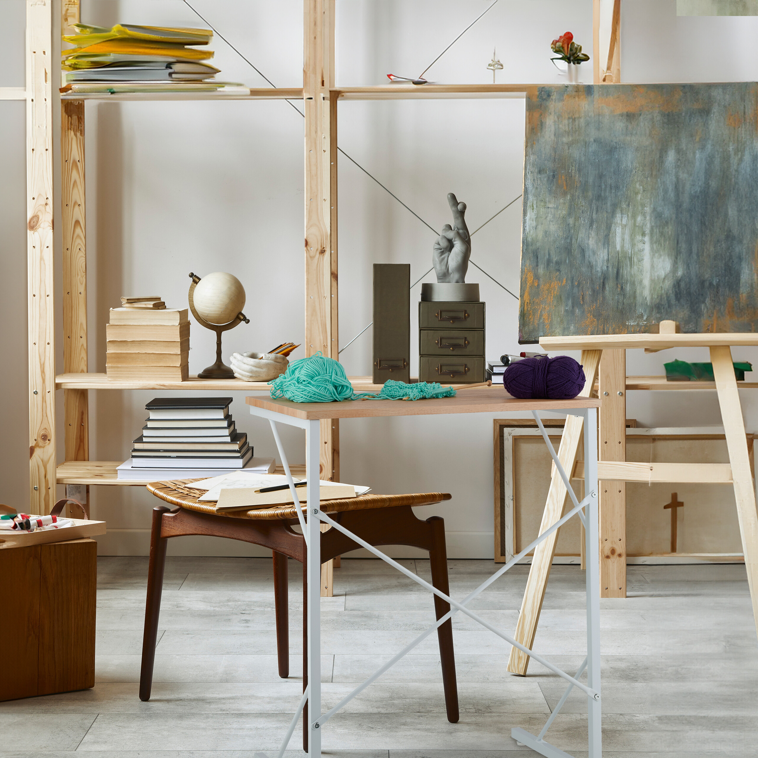 Atelierinterieur mit Holzböden, großem Regal voller Bücher, Staffelei mit Leinwand und kleinem Tisch mit Garn.