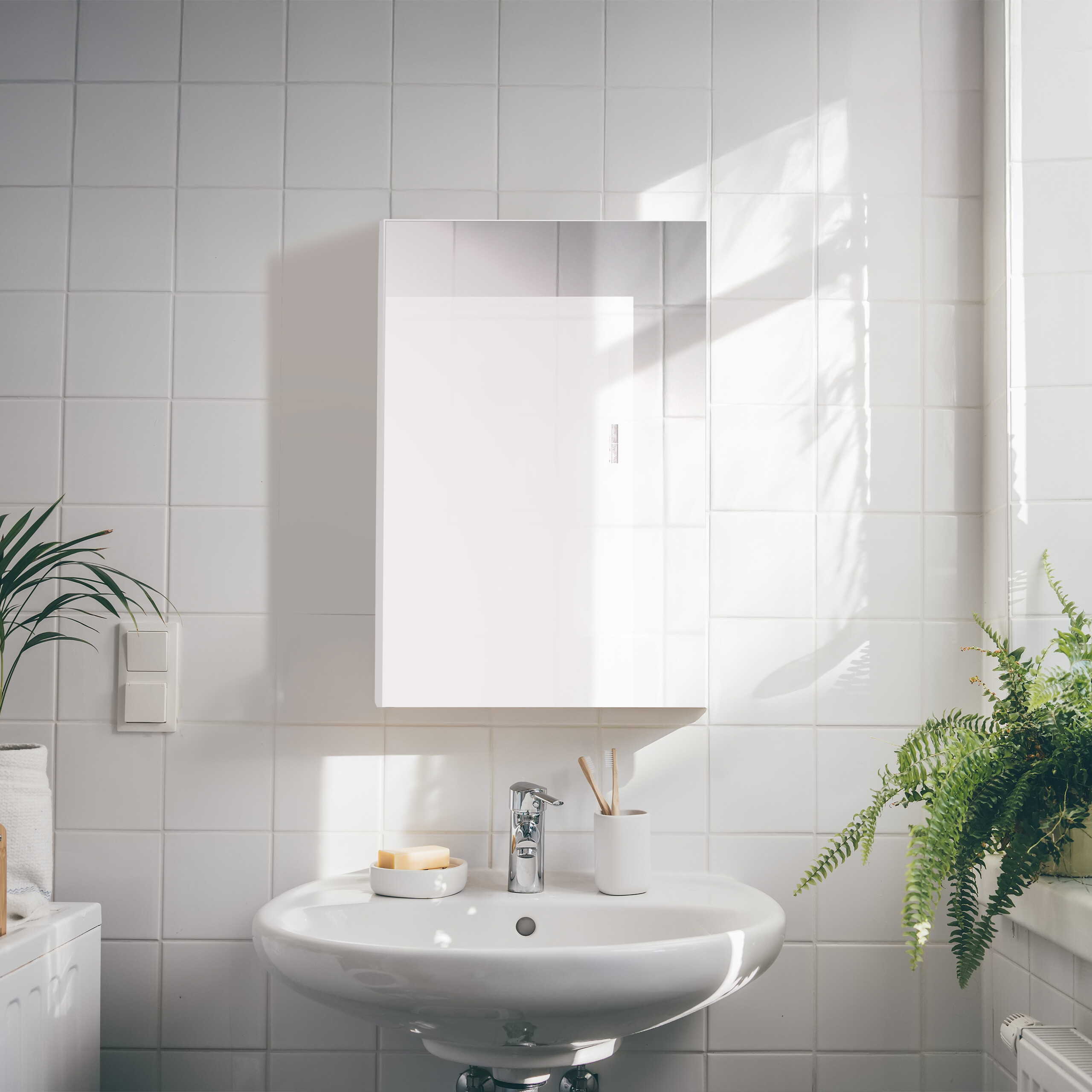 Ein weißes Waschbecken mit Armatur, Seife und Zahnbürsten unter einem Spiegelschrank an weißen Kacheln.
