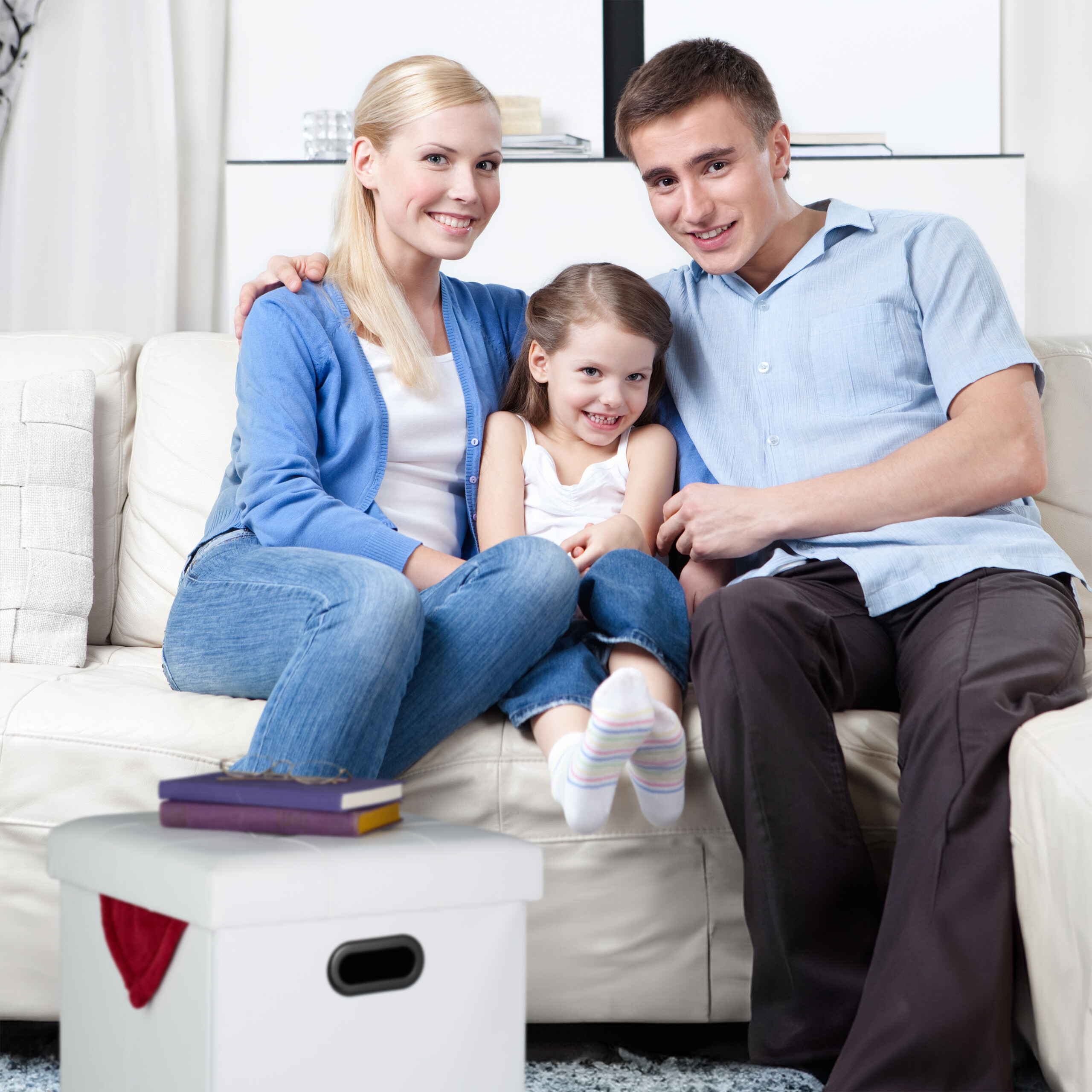 Eine lächelnde dreiköpfige Familie sitzt zusammen auf einem weißen Sofa in einem hellen Wohnzimmer.