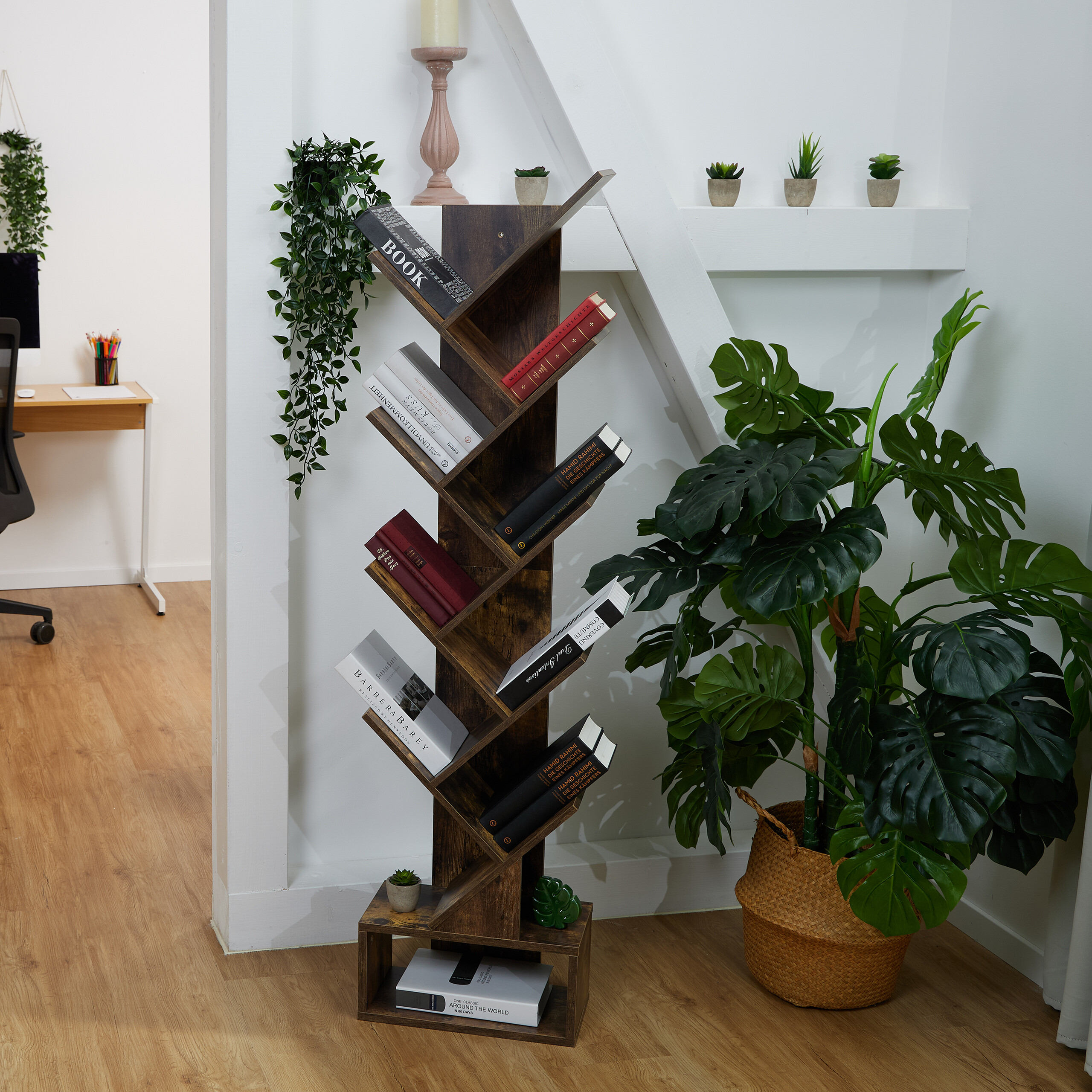 Ein holzfarbenes, baumförmiges Bücherregal steht an einer weißen Wand, gefüllt mit Büchern, umgeben von Pflanzen.