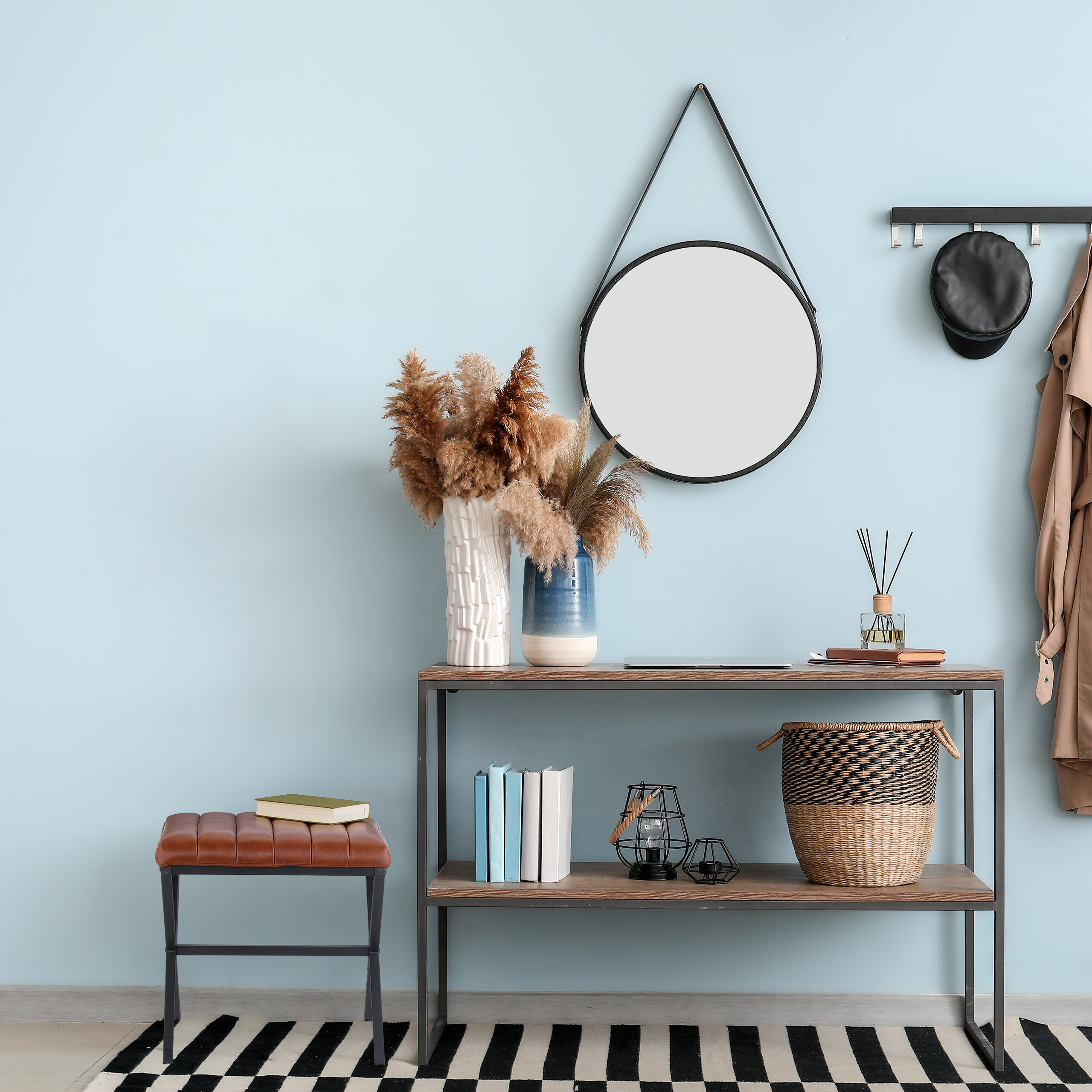 Ein Flur mit Konsole, rundem Spiegel, Hocker und Garderobe an hellblauer Wand auf gestreiftem Teppich.