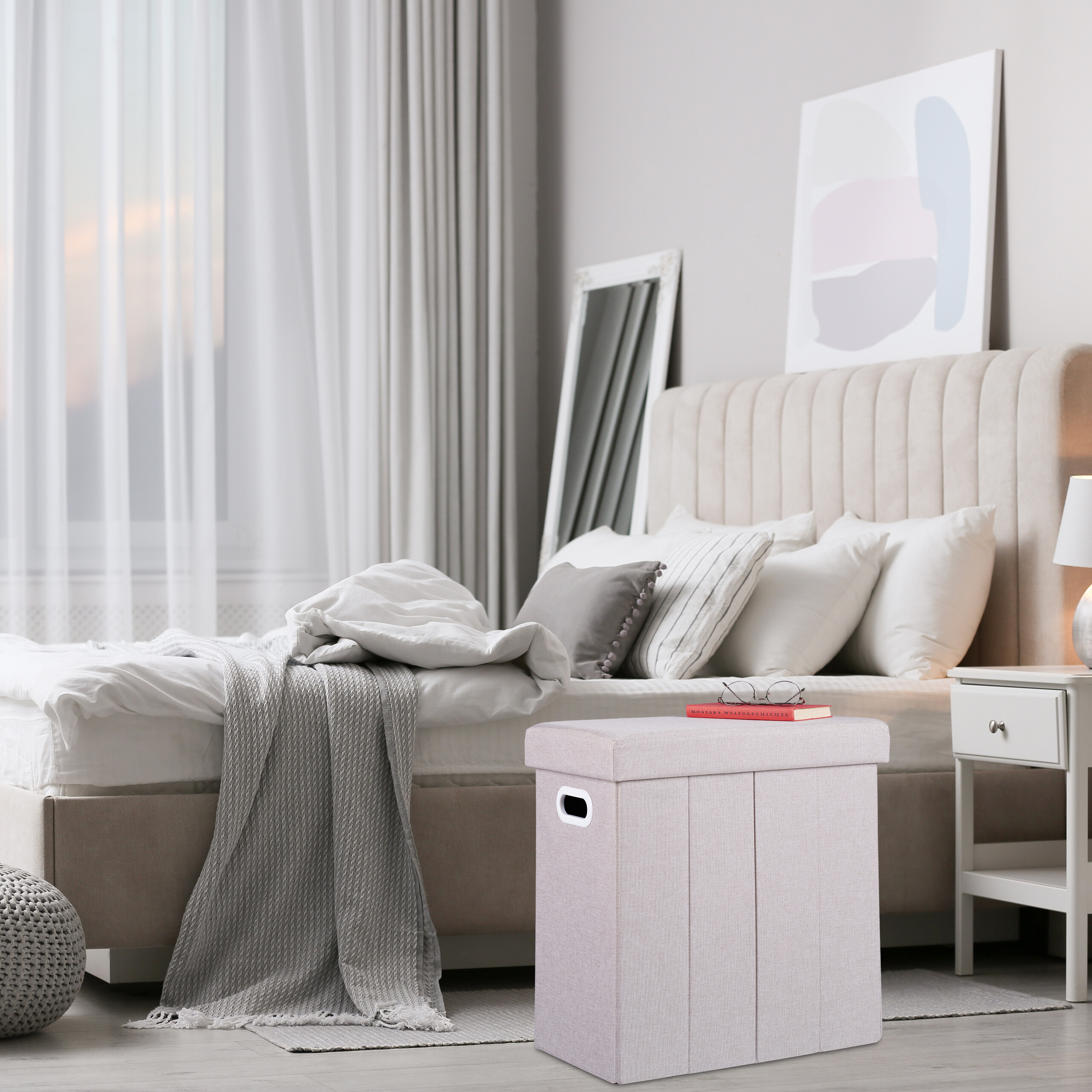 Ein gemütliches Schlafzimmer mit einem großen Bett, grauer Bettwäsche und einem hellen Aufbewahrungshocker im Vordergrund.