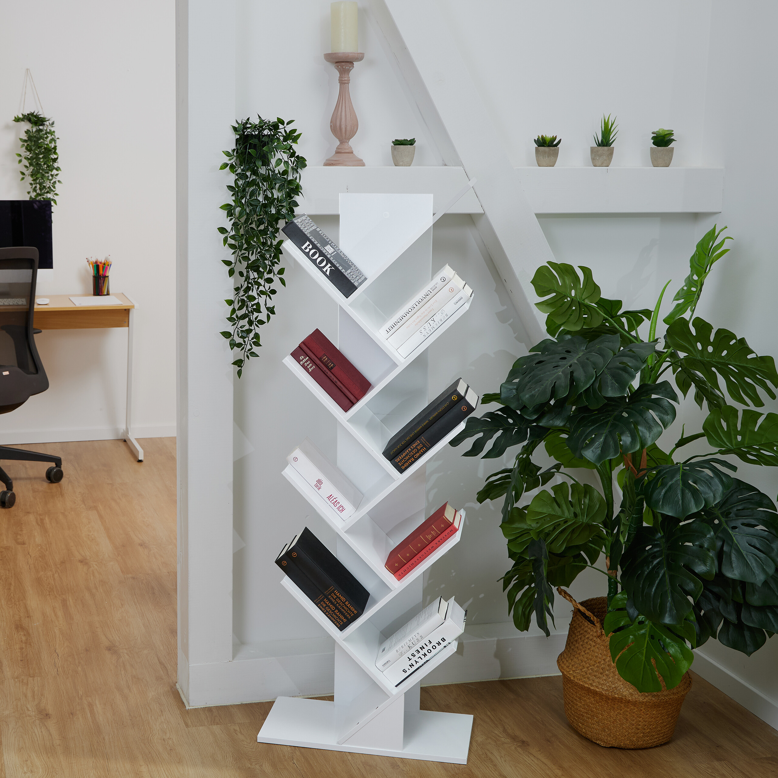 Ein weißes Bücherregal in Baumform mit Büchern steht neben einer großen Monstera-Pflanze in einem hellen Raum.