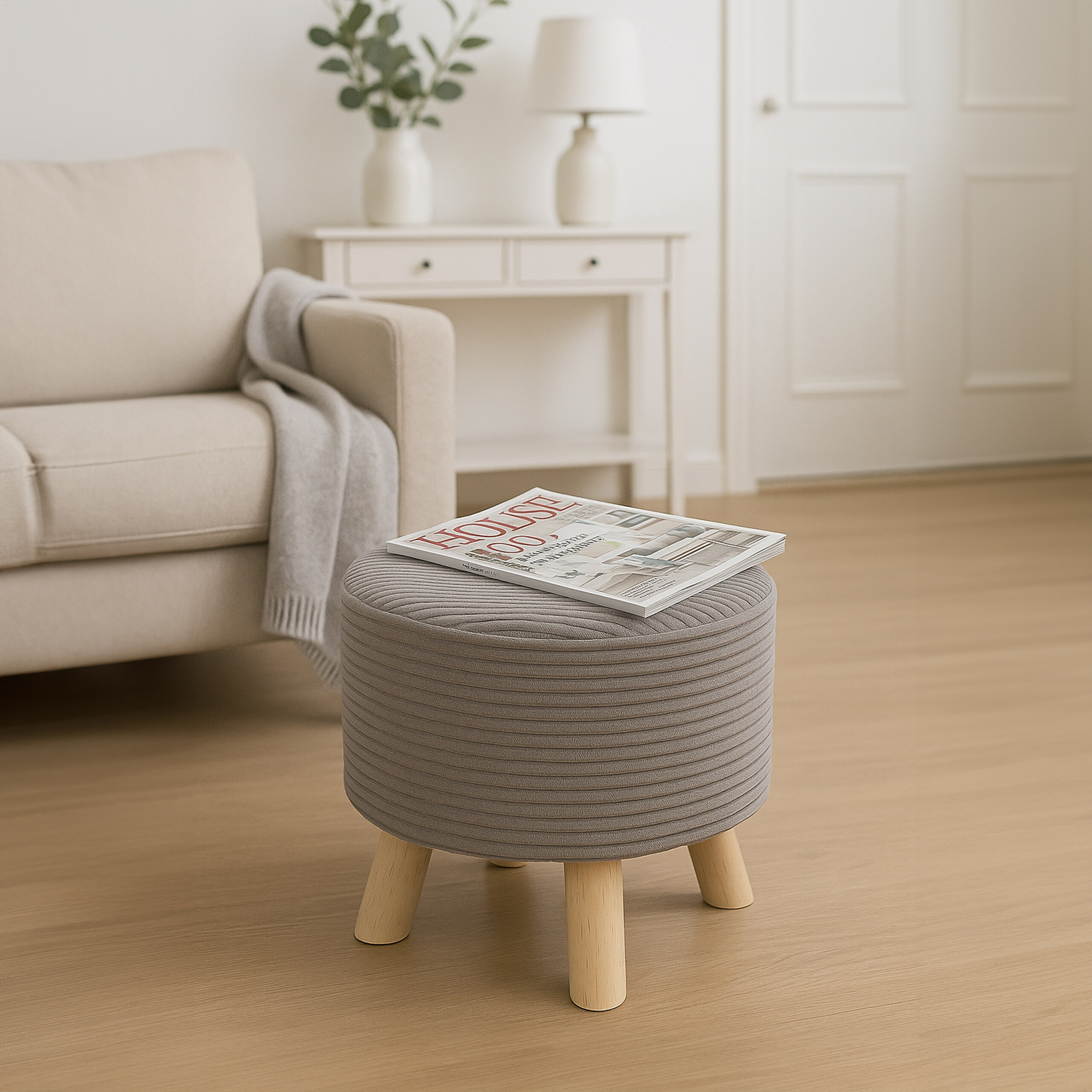 Ein grauer gerippter Hocker mit Holzbeinen steht auf einem Holzboden in einem hellen Raum mit einem Sofa im Hintergrund.