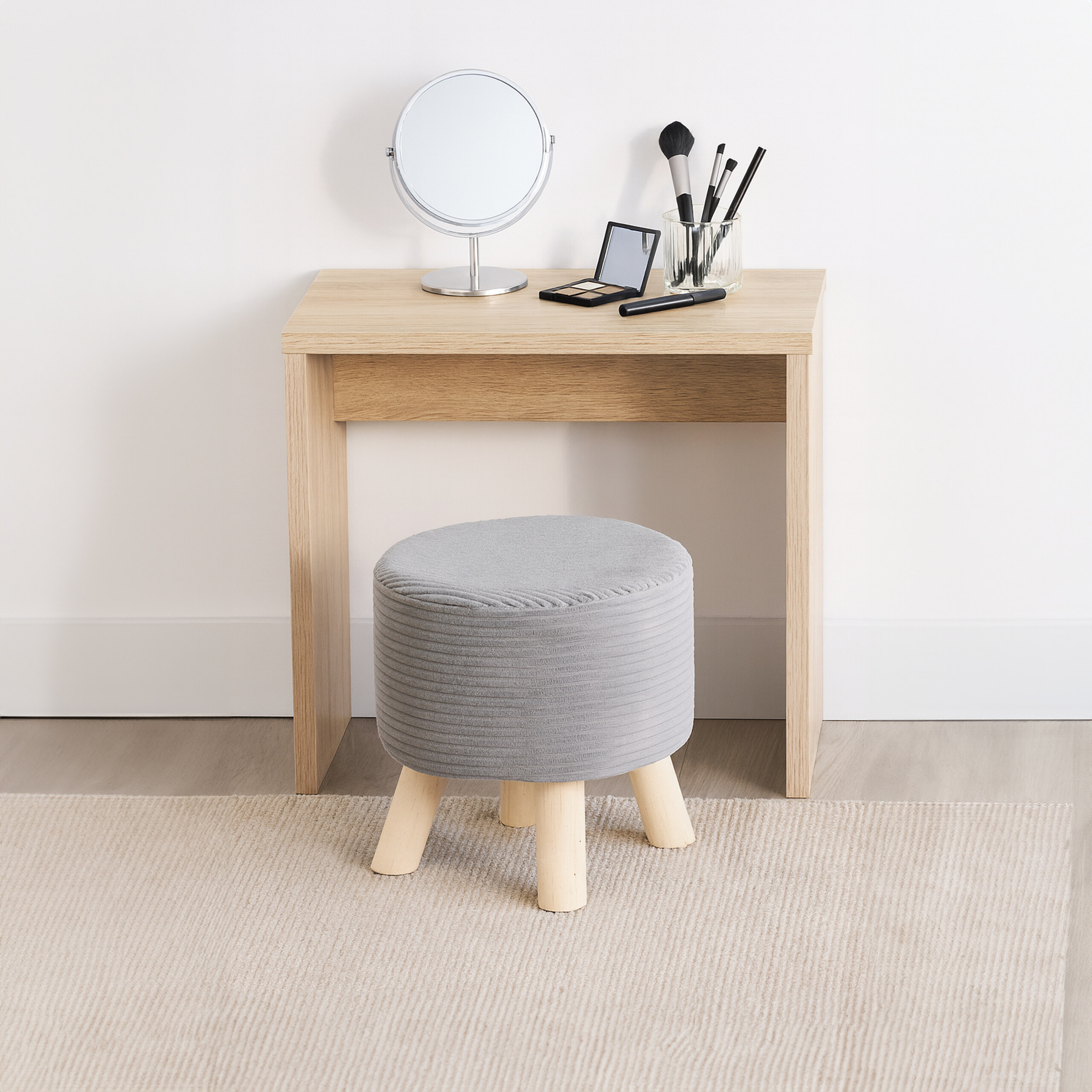 Ein Holztisch mit Spiegel, Make-up und Pinseln sowie ein grauer Hocker auf einem Teppich vor weißer Wand.