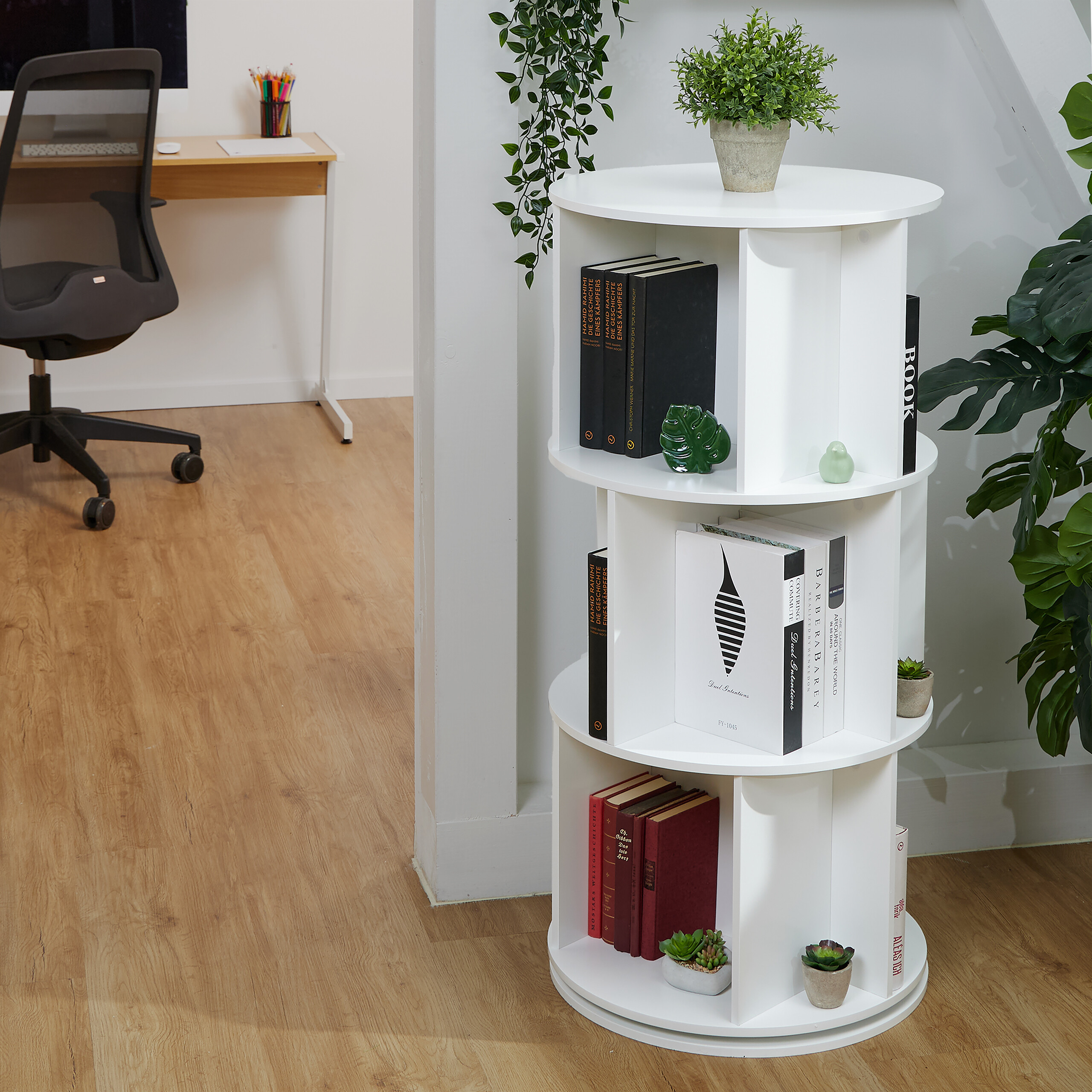 Weißes drehbares Bücherregal mit Büchern und Pflanzen im Innenbereich, im Hintergrund ein Schreibtisch.