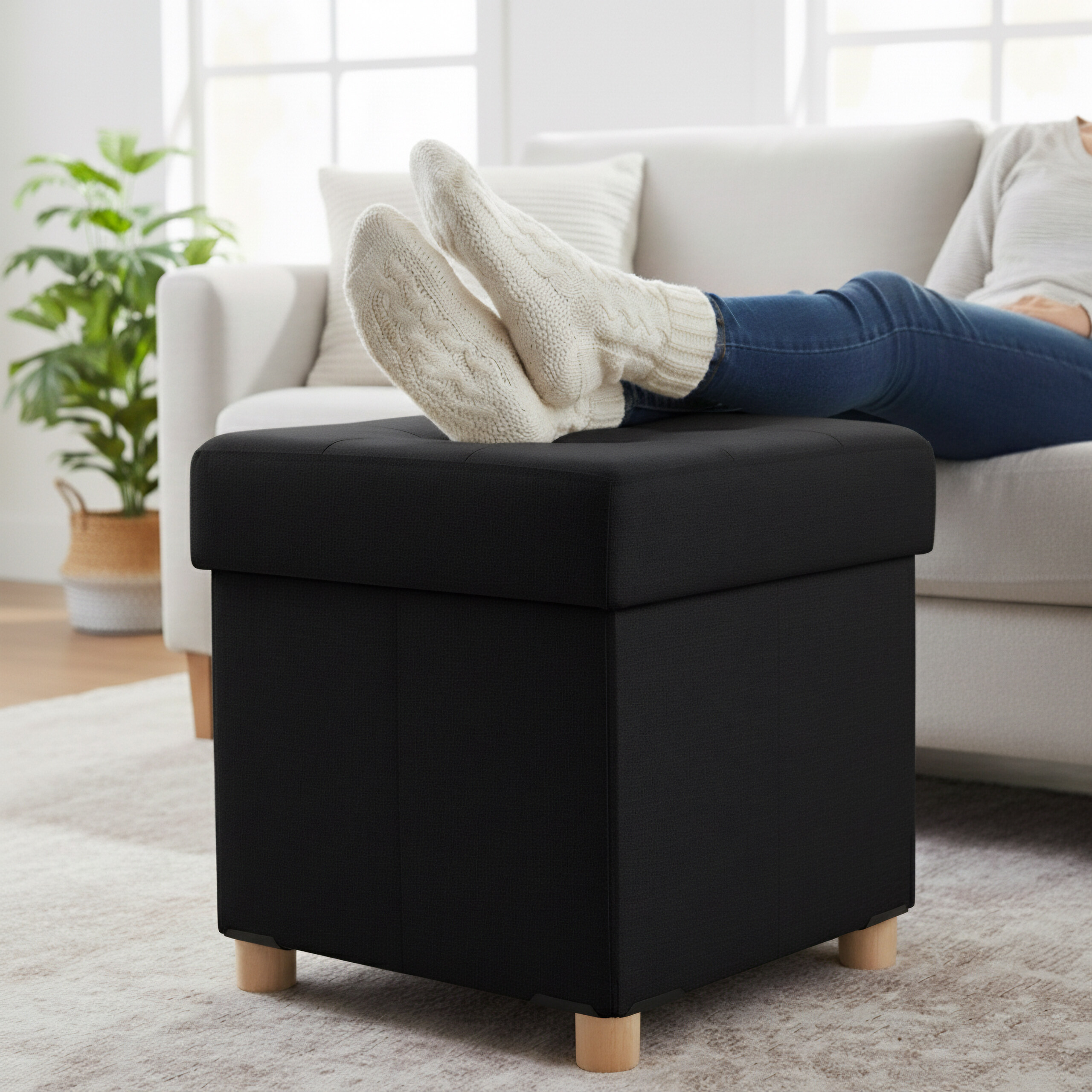 Eine Person sitzt auf einem Sofa und legt die Füße in dicken Socken auf einen schwarzen Hocker in einem hellen Zimmer.