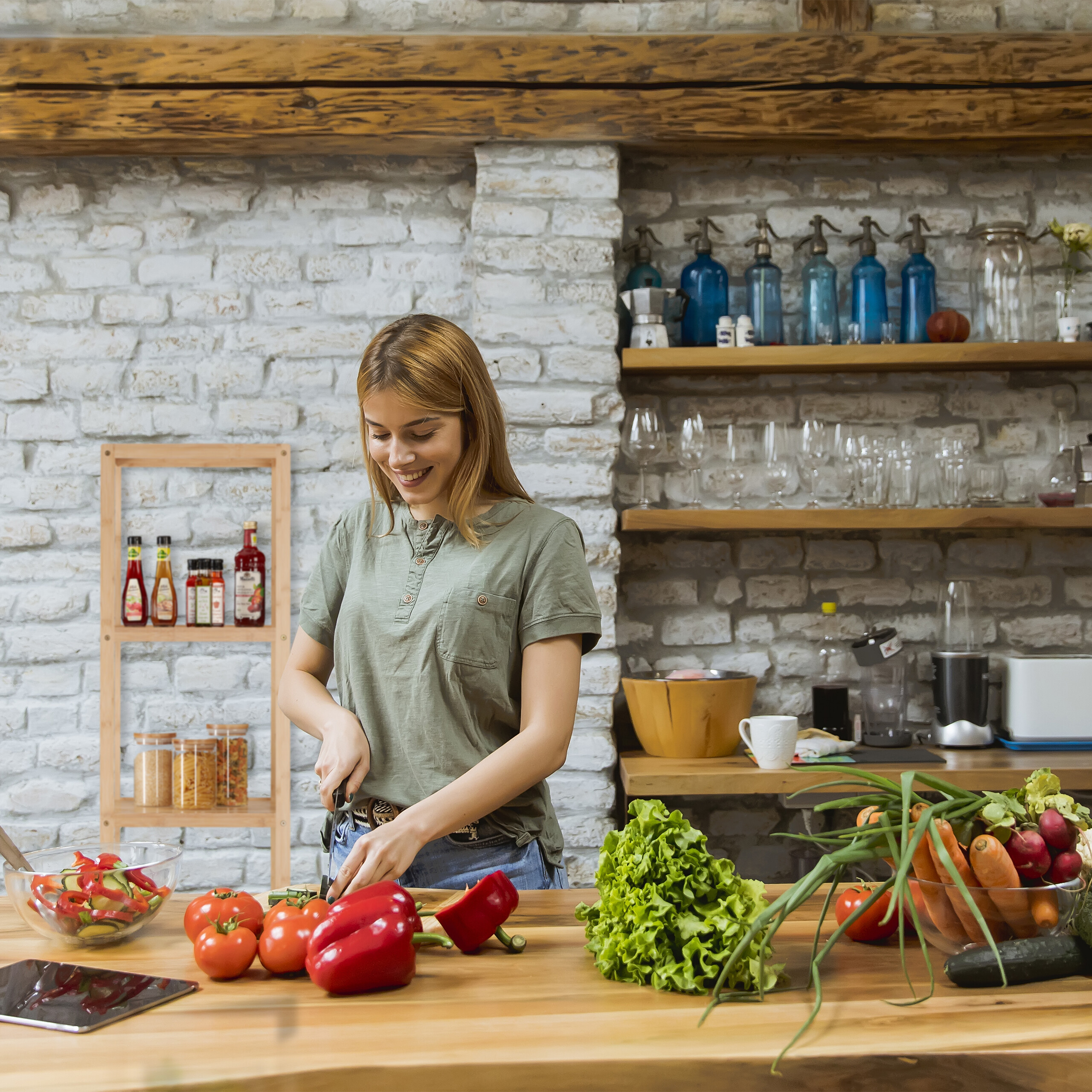 Eine Frau schneidet Gemüse in einer Küche mit Backsteinwand und Holzregalen voller Vorräte.