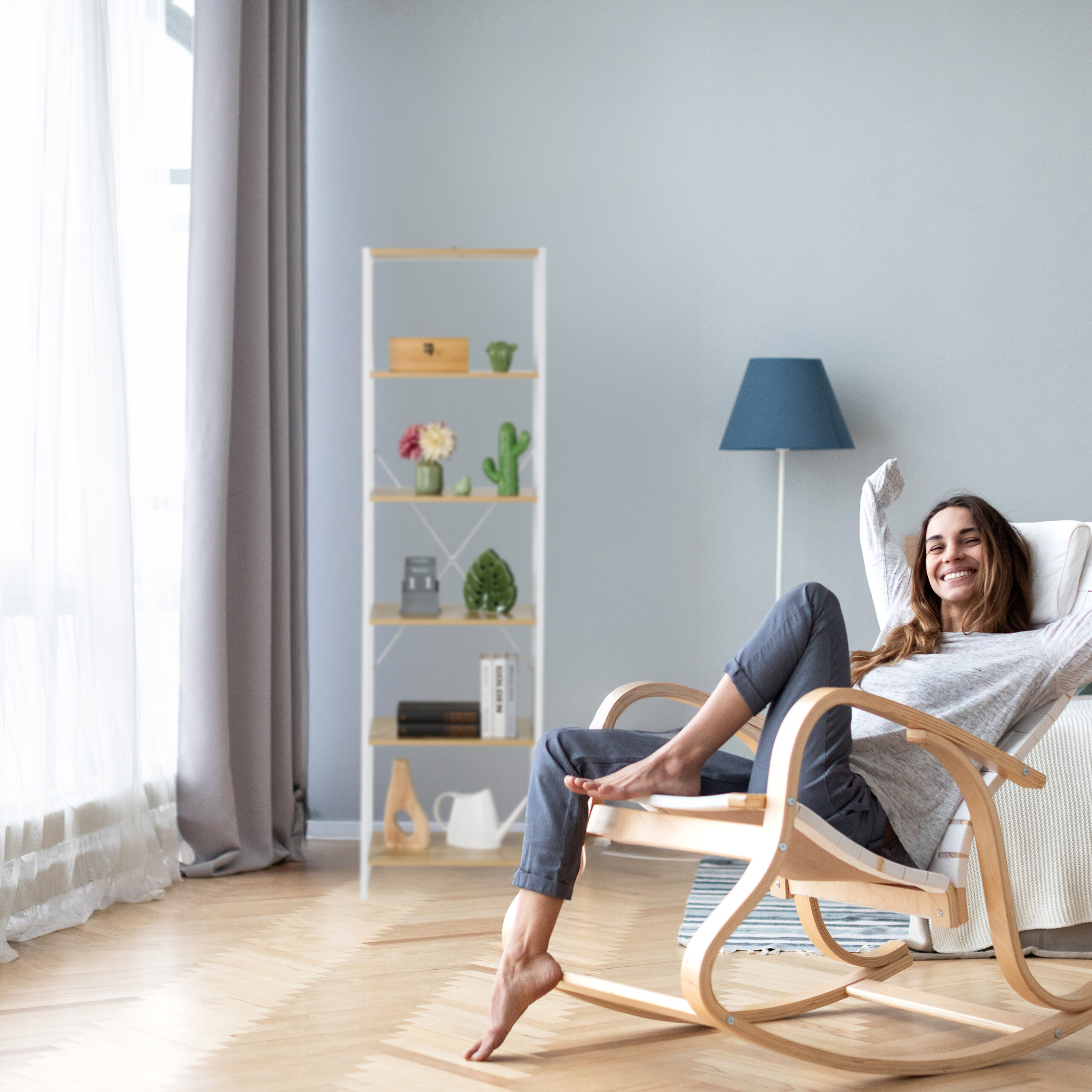 Eine lächelnde Frau sitzt in einem Holzschaukelstuhl in einem hellen, modern eingerichteten Wohnzimmer.